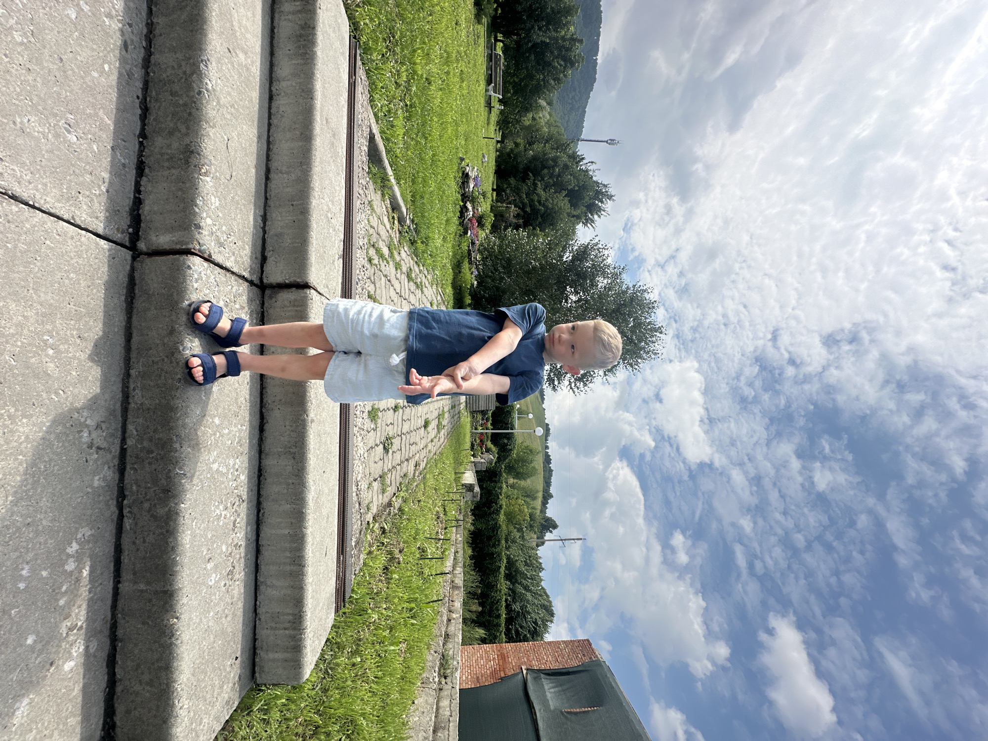 A young boy with blonde hair stands on concrete stairs outdoors. He is wearing a dark blue t-shirt and light blue shorts, with dark blue sandals on his feet. His arms are crossed in front of him, and he is looking to his right. The background shows a green grassy area with trees and hills in the distance. The sky is partly cloudy with a mix of white and gray clouds. The image appears to be taken in an outdoor park or recreational area. It is not possible to determine the specific location within Danilovka, Altai Krai, Russia, from the provided image.