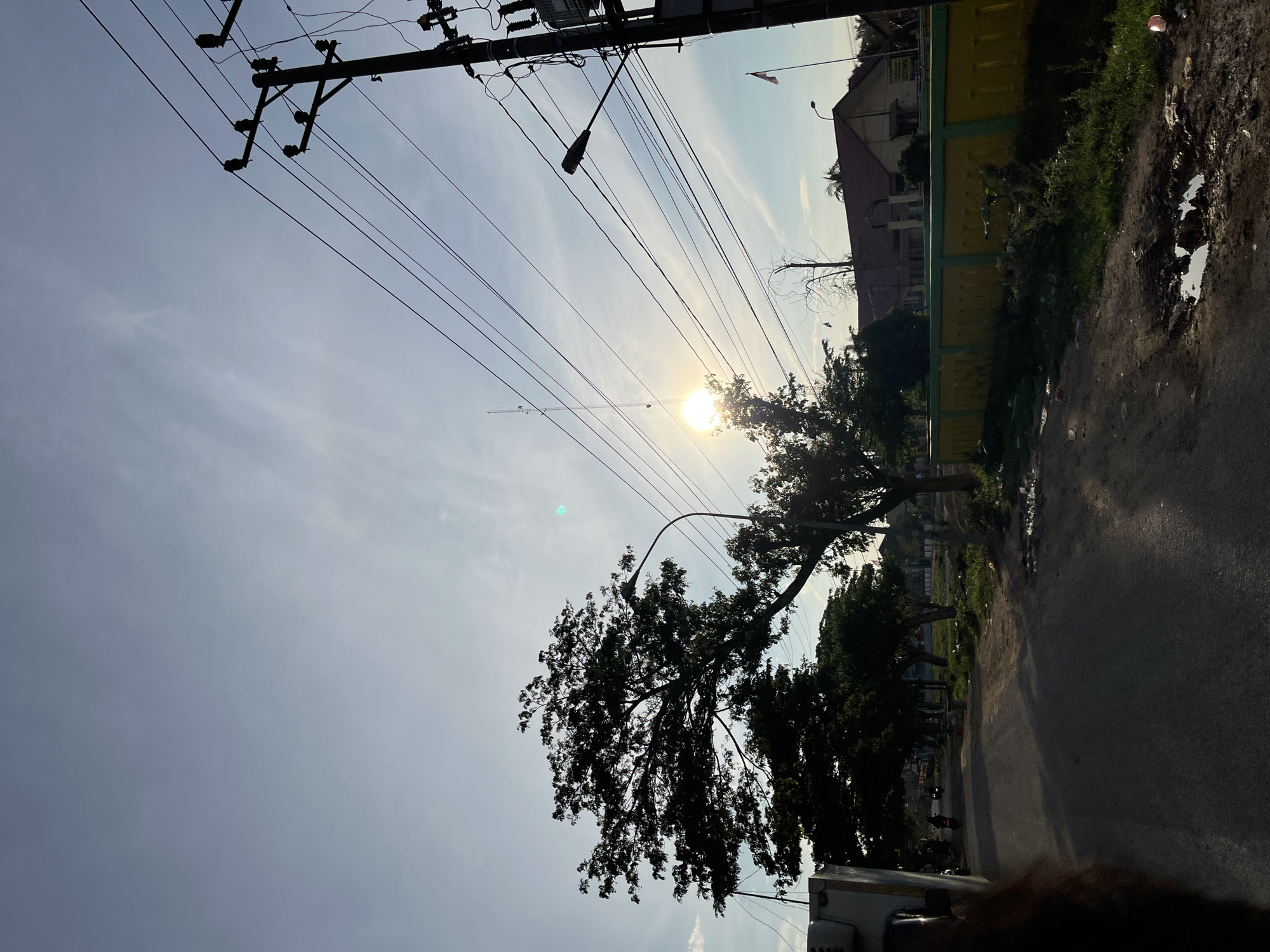 The image depicts a street scene at sunrise or sunset in Alurcucur, Indonesia. The sun is low in the sky, casting long shadows and creating a lens flare in the upper right quadrant of the frame. A large tree with a dense canopy dominates the left side of the image, silhouetted against the bright sky. Power lines crisscross the sky, supported by utility poles. A street lamp is visible on the right side of the road.
On the left side of the street, a vehicle, possibly a truck, is partially visible. Further down the road, a few people on motorcycles can be seen. A yellow and green fence runs along the right side of the street, with a building visible behind it. The road itself appears to be paved but has several potholes and puddles, indicating recent rain or poor road conditions. Vegetation, including grass and some smaller trees, grows along the roadside. A flagpole with a white and red flag, presumably the Indonesian flag, is visible in the distance on the right. The overall impression is of a somewhat rural or suburban street in Indonesia during a transitional time of day.