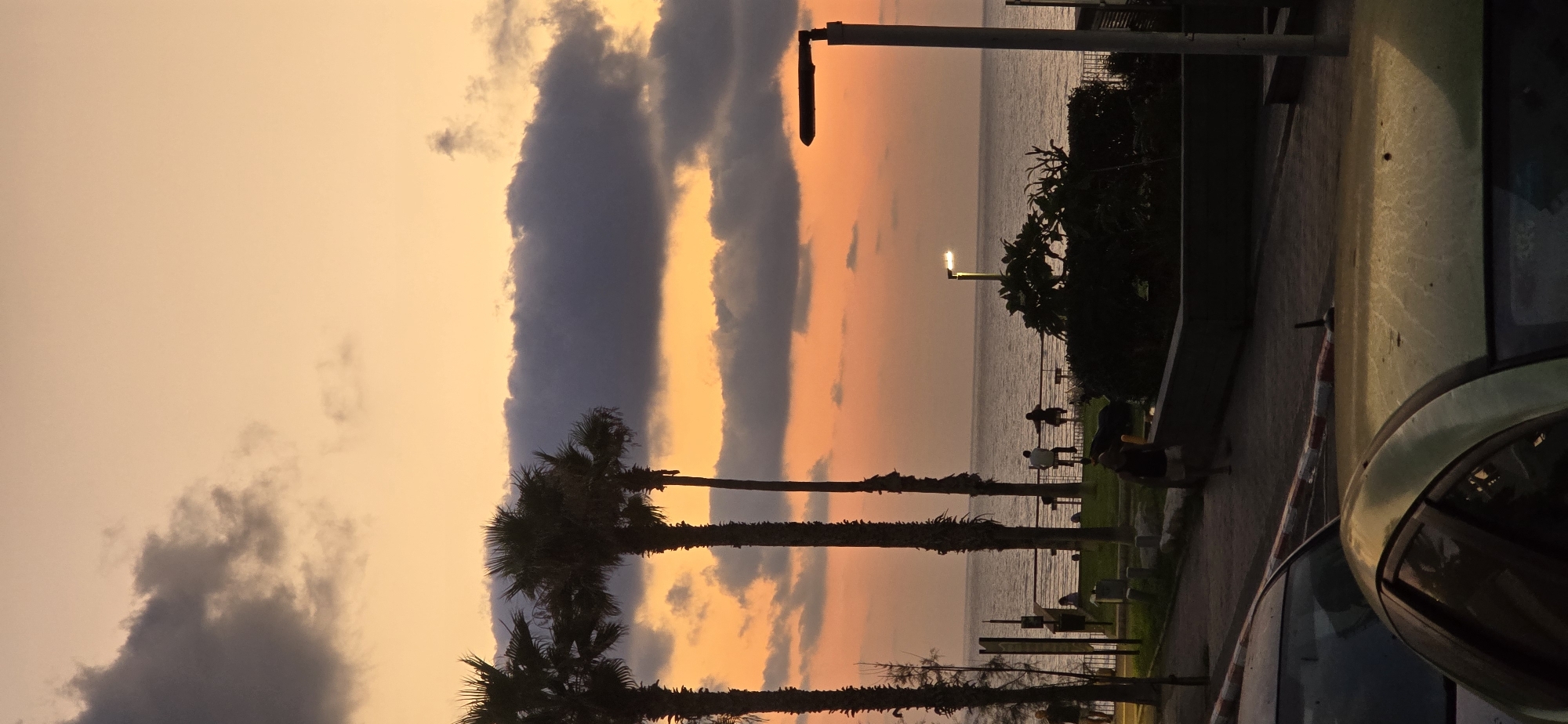 The image depicts a scenic sunset over the Mediterranean Sea in Bat Yam, Israel. Three tall palm trees stand silhouetted against the colorful sky, which transitions from a soft orange to a lighter yellow hue. Dark, wispy clouds are scattered across the horizon. Along the coastline, a pathway lined with a metal railing is visible, with several individuals seen standing or sitting. To the right, a modern street lamp with a single light fixture illuminates the scene. The foreground shows a street with parked cars and a pedestrian area. The overall atmosphere is tranquil and serene, capturing the beauty of a coastal sunset.