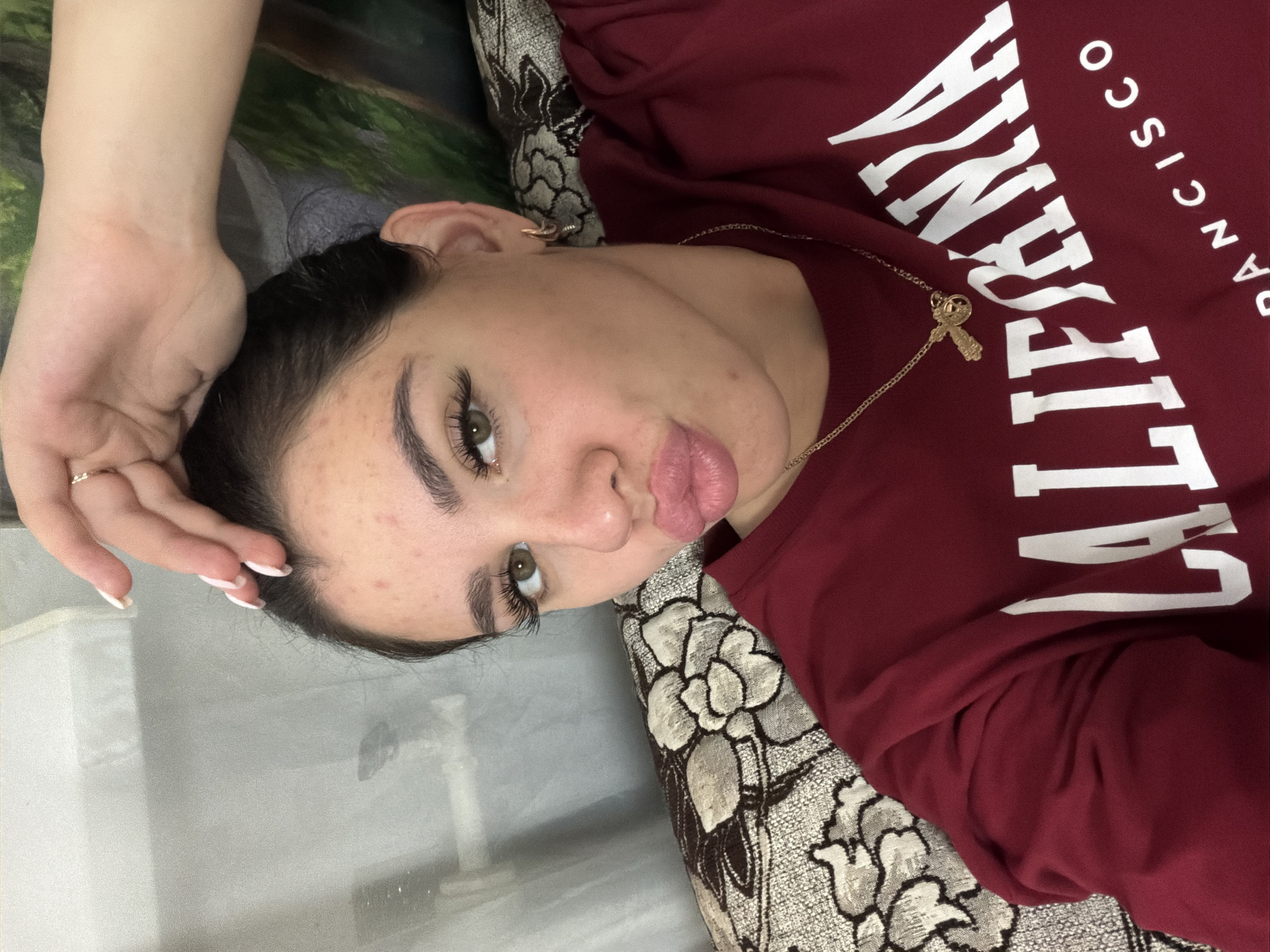 A young woman with dark hair is captured in a close-up, selfie-style shot, resting on what appears to be a couch with a floral-patterned cushion. She is wearing a maroon sweatshirt with "CALIFORNIA" and "FRANCISCO" printed in white block letters. A thin gold chain with a cross pendant is visible around her neck. Her right hand is raised, fingers touching her forehead, and she is looking directly at the camera with a pouty expression. The background is blurred, suggesting an indoor setting. No specific location in the city of Фоки, Russia, can be determined from the image.