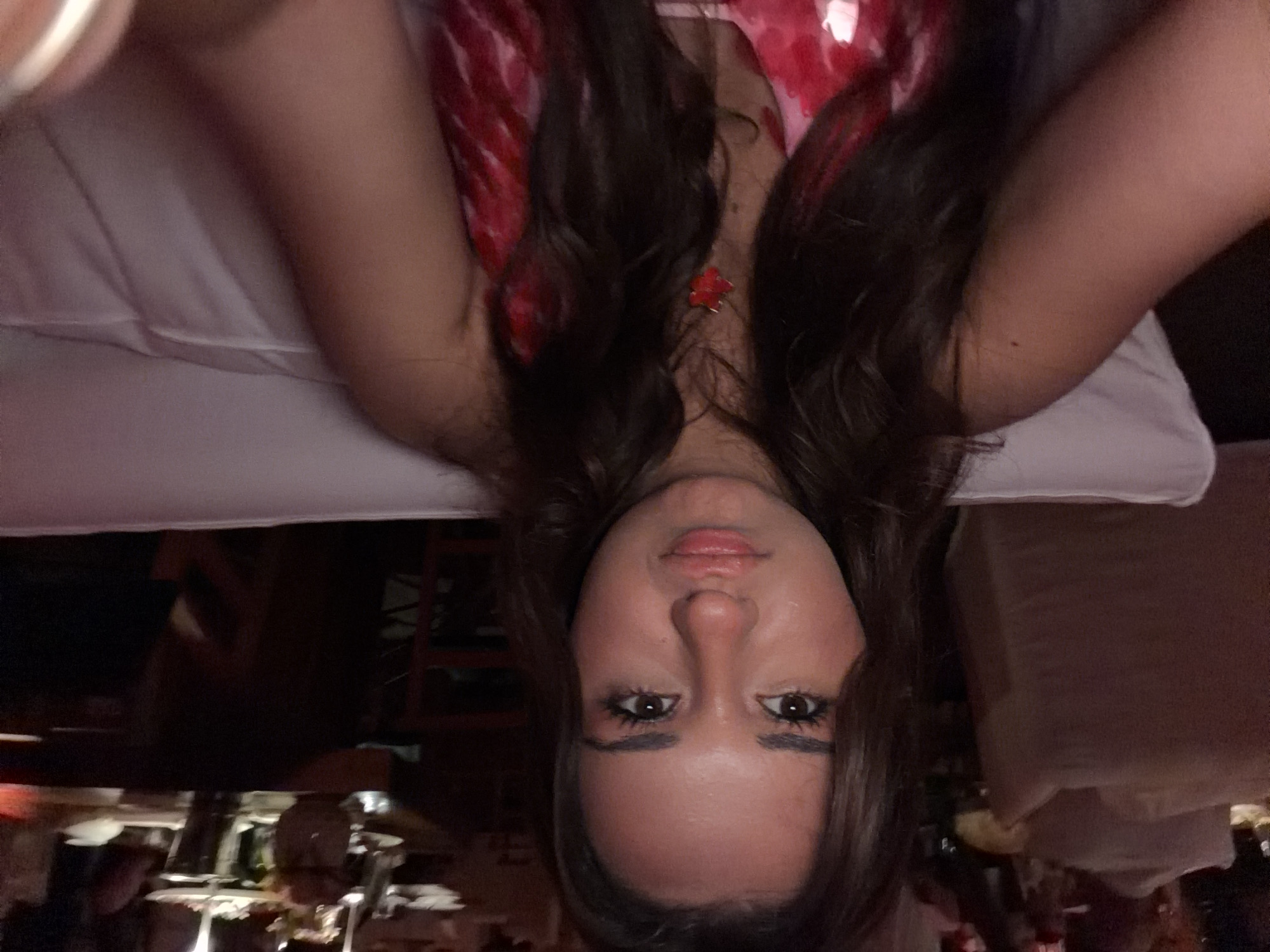 A young woman with long, dark, wavy hair and brown eyes is in the foreground of the image, looking directly at the camera. She has full lips and is wearing a red and pink patterned dress. A red pendant necklace is visible around her neck. She appears to be seated on a white cushioned sofa in a dimly lit restaurant or lounge. In the background, blurred figures and tables with place settings can be seen, suggesting a social gathering. The environment appears to be an indoor establishment, possibly a bar or restaurant in Yerevan, Armenia, given the information provided. The lighting is warm and atmospheric, with some sources of light in the background creating a soft glow.