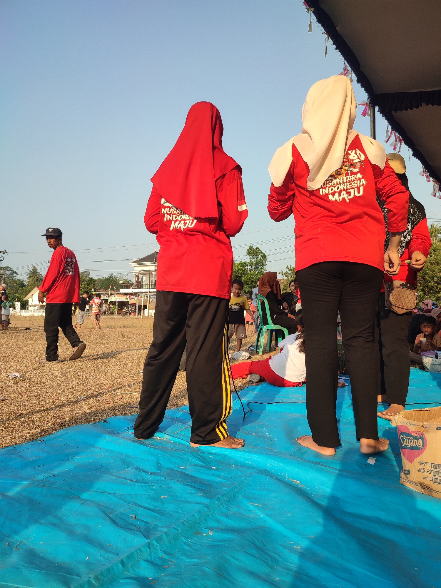 The image depicts an outdoor scene in Nusa Jaya, Indonesia, likely a community event or festival, judging by the attire and activities. The foreground is dominated by a bright blue tarp spread on dry, golden grass. Several people are visible, primarily wearing red shirts with Indonesian text, suggesting a coordinated group. Two individuals in the foreground are seen from behind, both wearing red shirts and black pants; one has a red hijab, and the other wears a light-colored hijab. Their bare feet are visible on the blue tarp. To the left, a man in a red shirt and black pants, wearing a black baseball cap, walks away from the camera, also with bare feet. In the middle ground, other individuals are gathered, some sitting on chairs and on the ground, including children. A woman in a white shirt and red pants is lying on the blue tarp. The background shows a typical Indonesian village setting with houses, trees, and utility poles under a clear, bright sky. A cardboard box with visible Indonesian text, "Sayang," is placed on the blue tarp on the right side of the frame. The overall impression is one of an informal gathering in a public space.