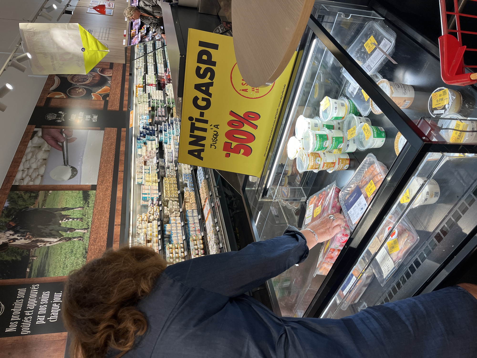 The image depicts the interior of a supermarket in Andrézieux-Bouthéon, France, likely during daylight hours. A woman with reddish-brown curly hair, wearing a dark blue shirt, is seen from the back, reaching into a refrigerated display case. The display case is filled with various food products, including packaged cheeses, yogurts, and possibly seafood. Many of these items have yellow or red discount stickers, indicating a sale.
A prominent yellow sign with bold black text reads "ANTI-GASPI JUSQU'À -50%", advertising a "no waste" initiative with a 50% discount. The sign also mentions "Offre valable sur tous les produits stickés ANTIGASPI, remise déjà effectuée," meaning the offer is valid on all products marked ANTIGASPI, with the discount already applied.
Further in the background, more shelves stocked with dairy products are visible, along with other shoppers browsing. A large mural on the wall features images of cows and agricultural scenery, alongside text in French that translates to "Our products are tasted and approved by us every day." The overall atmosphere suggests a typical grocery shopping experience, with a focus on discounted items. The lighting appears to be artificial, typical of an indoor retail environment.