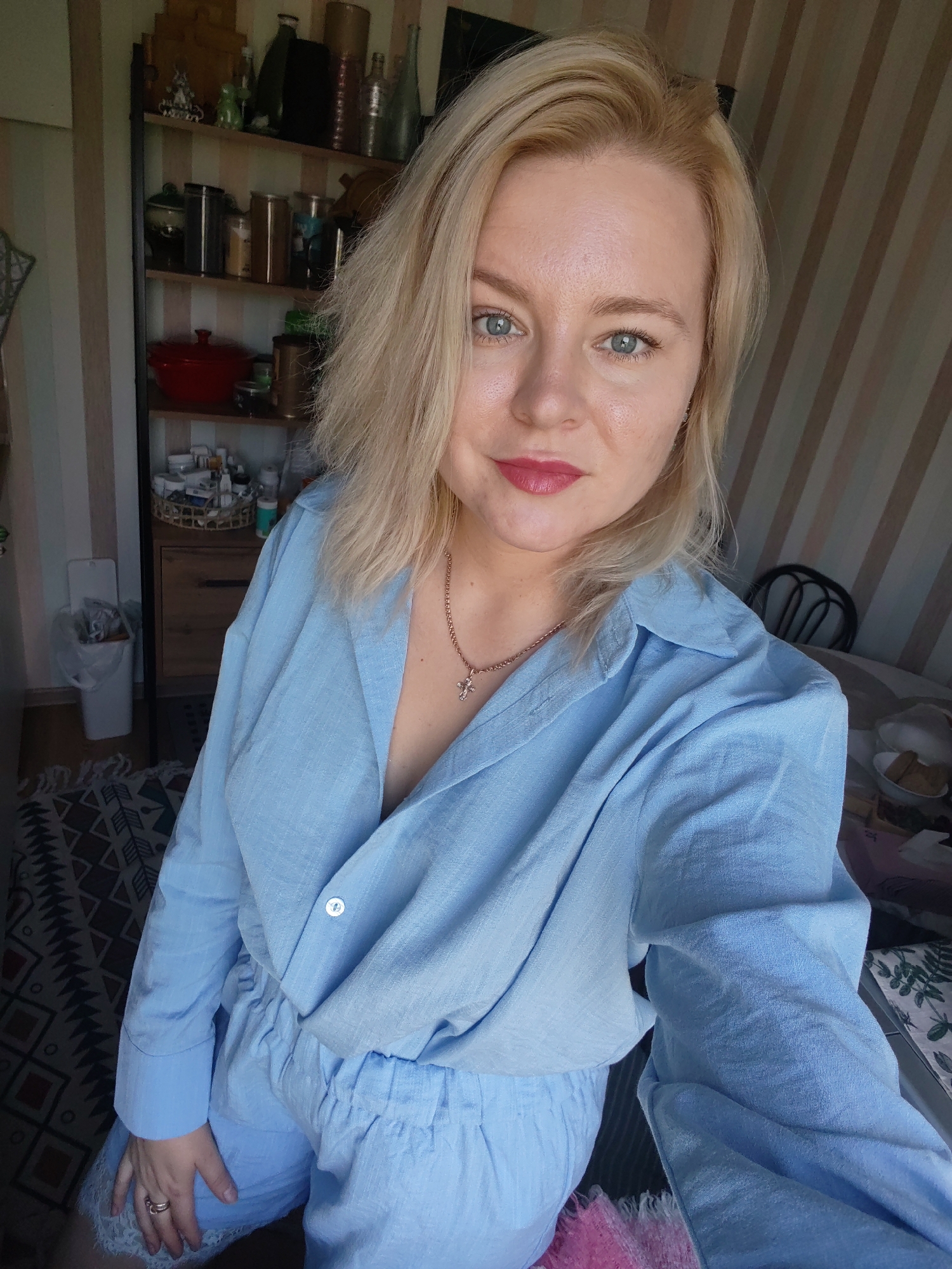 A blonde woman with blue eyes and red lipstick is taking a selfie indoors. She is wearing a light blue collared shirt and light blue shorts. She has a necklace with a cross pendant. In the background, there are shelves with various items, including jars, bottles, and a red pot. There is also a trash can, a rug, and a table with some food on it. The walls are striped. The scene appears to be in a domestic setting in the city of Izhevsk, Russia.