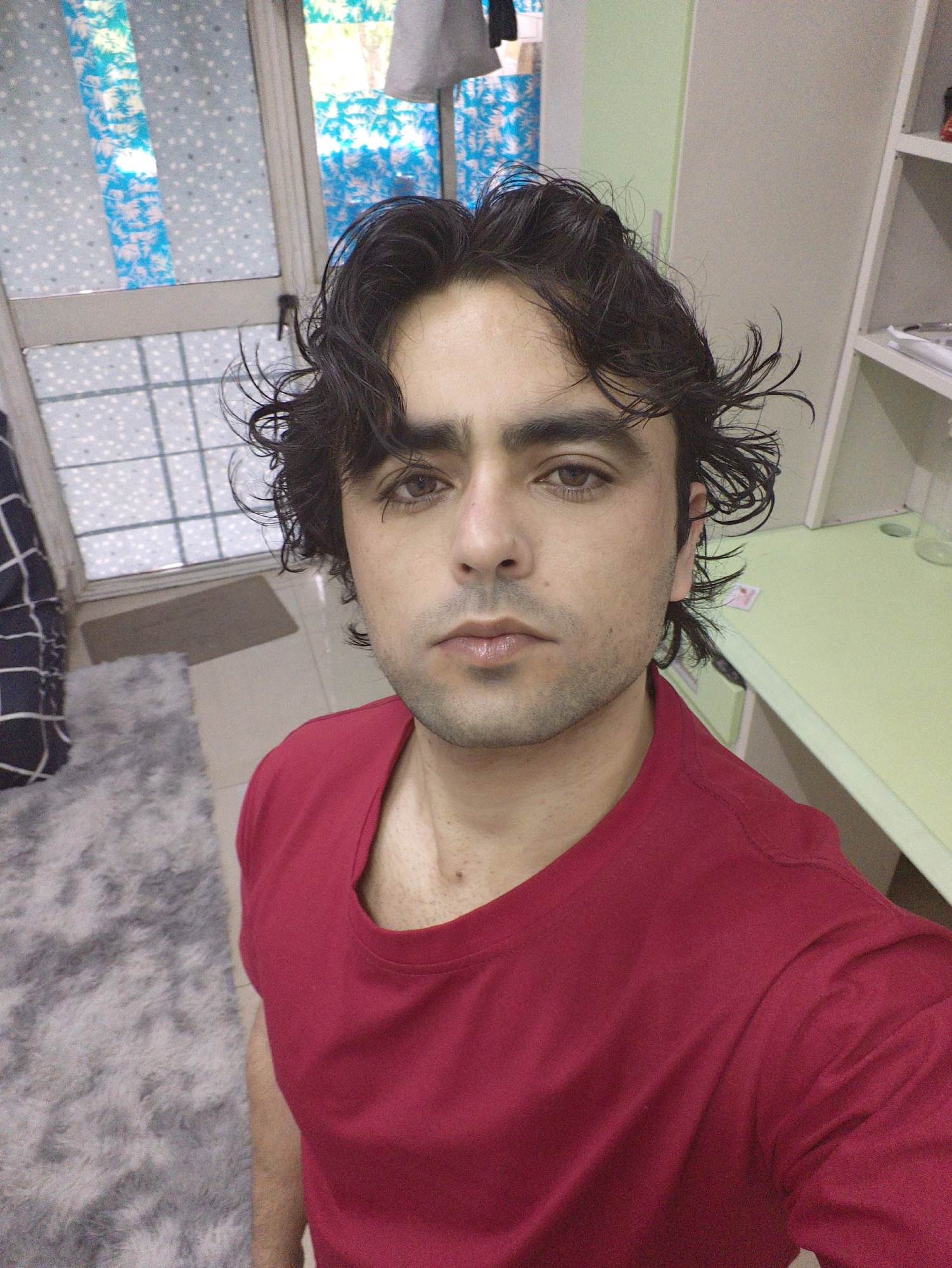A young man with dark, wavy, and somewhat dishevelled hair takes a selfie indoors. He has dark eyes, light stubble, and a neutral expression, looking directly at the camera. He is wearing a plain red short-sleeved T-shirt.
The setting appears to be a room within a residence, possibly a bedroom or study, located in Chongqing High-tech Zone, China. To the man's left, a grey, shaggy rug covers a tiled floor, and a darker, smaller brown mat is positioned near a glass-paneled door and window. Both the window and the upper part of the door are covered with a semi-transparent blue and white patterned film, which diffuses the natural light coming from outside. Hints of bright light and possibly distant greenery are visible through the film or its less dense areas, suggesting it is daytime.
To the man's right, a light green desk surface is visible, with white shelving units above and beside it. A few indistinct items, including what looks like a small glass or cup and a white card, are on the desk. The wall behind the shelving is a light, muted green. Behind the man's left shoulder, a dark patterned (possibly checkered) blanket or fabric is visible. No discernible text is visible in the image.