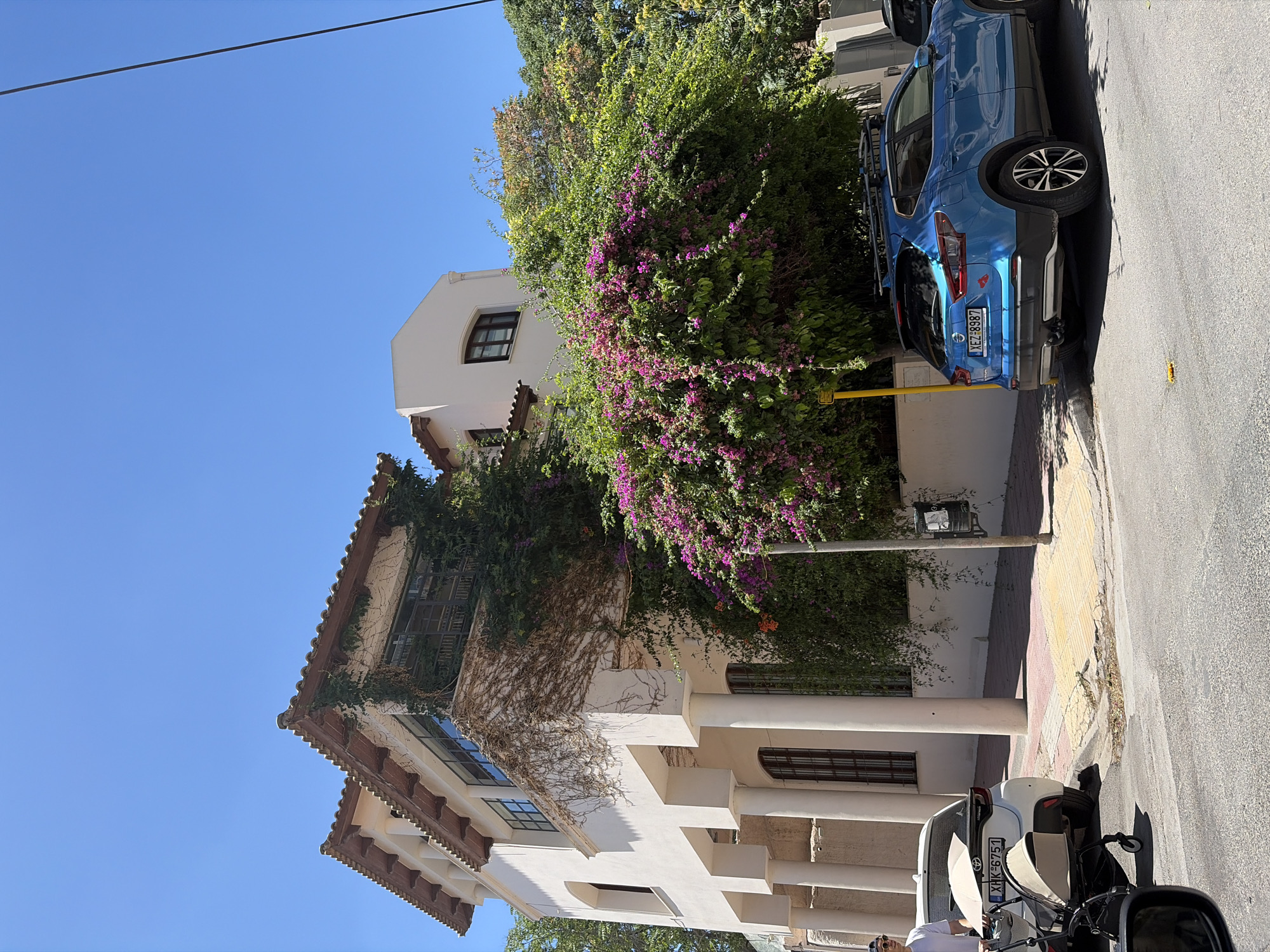 A bright, sunny daytime exterior shot captures a multi-story stucco building with a terracotta tile roof. The building features a prominent facade with a series of large, rounded columns on the lower level and a cantilevered upper floor with a balcony draped in lush green vines and vibrant purple bougainvillea. The building is flanked by dense foliage and trees. In the foreground, a blue SUV with a roof rack is parked on the street, facing away from the camera. To the left of the SUV, a white hatchback is parked, with a person pushing a stroller beside it. The street is paved, and a sidewalk with yellow and red patterned tiles runs along the side of the building. The sky is a clear, vibrant blue.
