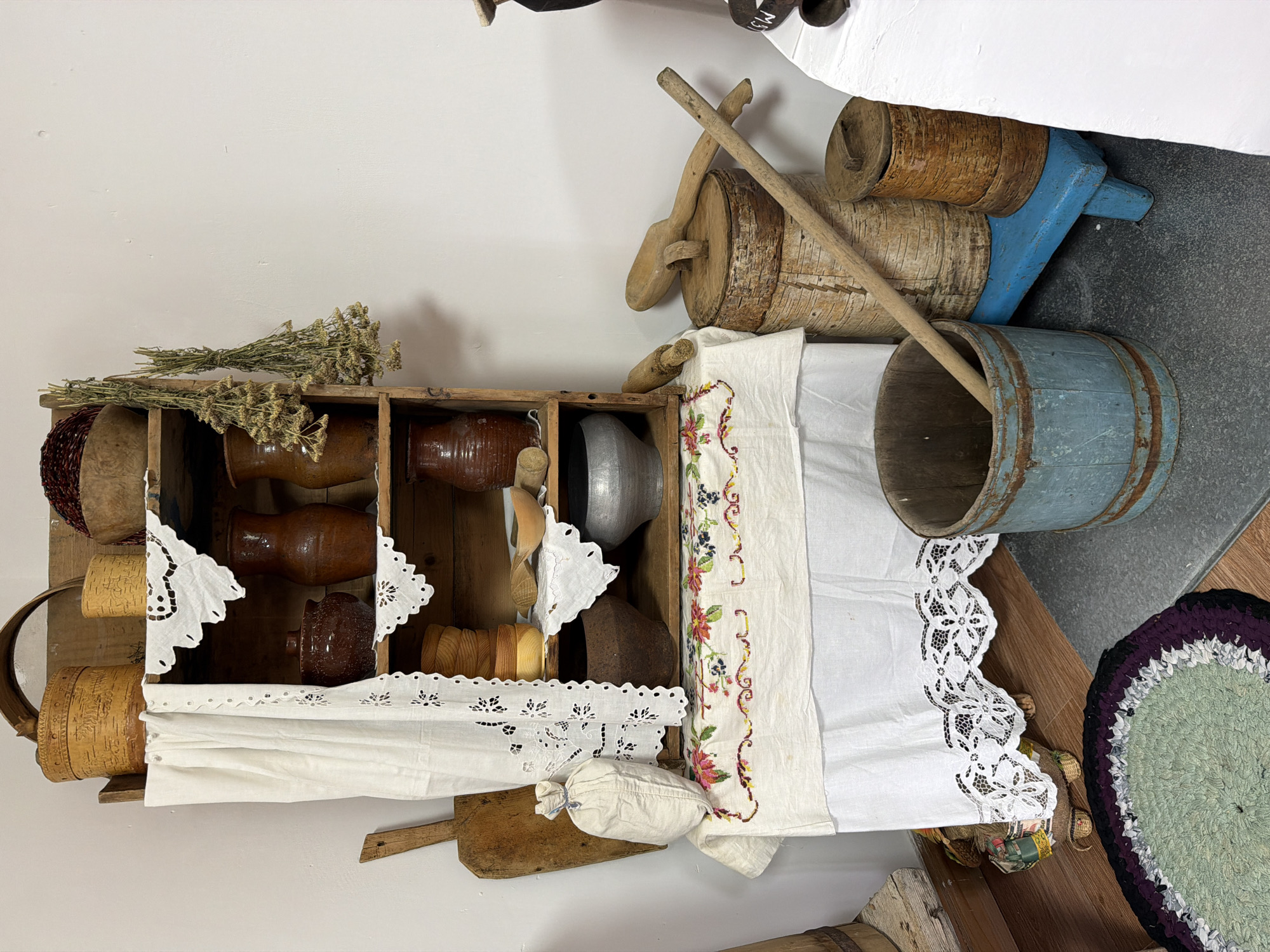 This is a forensic, matter-of-fact description of a staged domestic interior scene, likely in Ust'-Pyzas, Russia, depicting a collection of rustic, traditional household items. The scene is lit by ambient light, creating shadows that add depth to the objects.
The central focus is a wooden shelving unit filled with various earthenware and wooden vessels. On the top shelf, a large wooden bowl contains dark red material, possibly dried berries or grain. Next to it are two birch bark containers, one larger than the other. A wooden-rimmed basket is positioned to the left.
The shelves below are adorned with white lace-trimmed cloths. Earthenware jugs and pots of various sizes and shapes are displayed prominently, some with a glossy finish, others matte. There are also stacks of small wooden bowls and cups, and a larger metal pot on one of the lower shelves. A rolled-up white cloth hangs on the left side of the shelving unit, partially obscuring the view of some items.
To the left of the shelving unit, a wooden paddle or board leans against the wall. In front of the shelving unit, a small white cloth bag is tied at the top. Below the shelving unit, a white embroidered tablecloth with floral and berry motifs is draped.
On the floor in front of the shelving unit and to the right, several other items are arranged. A light blue wooden bucket with metal banding contains a long wooden pole. Behind this bucket are two larger wooden cylindrical containers, possibly butter churns or storage containers, with one having a wooden lid and a large wooden spoon resting on it. A small blue wooden stool supports one of these cylindrical containers. To the far right, a white surface, possibly a table or counter, is visible, with some dark objects on it. A circular rag rug in shades of green, purple, and black is situated on the floor to the lower left.
The overall impression is one of a carefully curated display of historical or traditional household artifacts, evoking a sense of rural life. There are no people present in the scene, and no actions or interactions are depicted. The environment appears to be an indoor setting with a plain white wall in the background and wooden flooring.