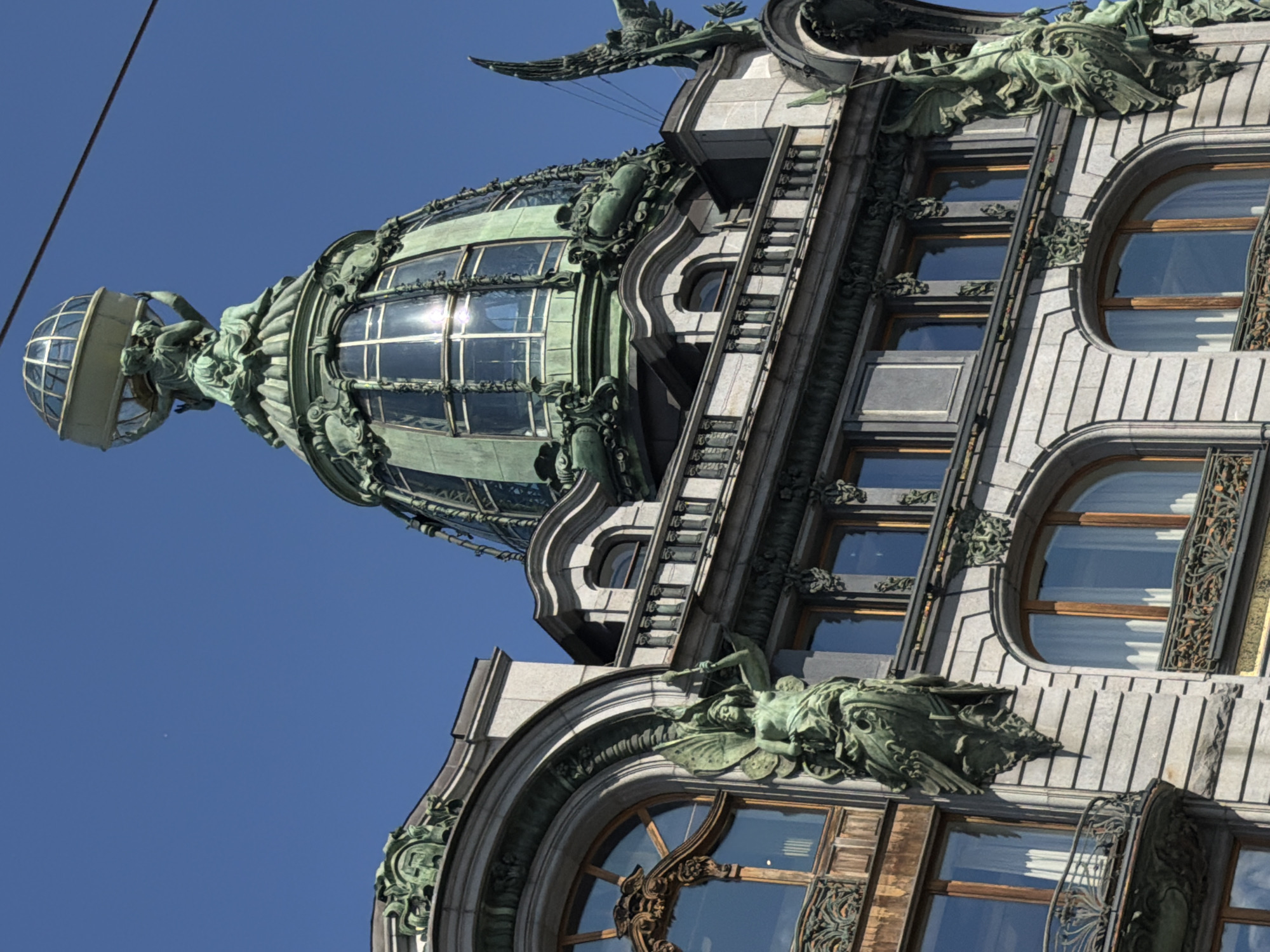 The image shows a detailed view of the Singer House, also known as the Dom Knigi, located on Nevsky Prospekt in Saint Petersburg, Russia. The iconic building features a striking green copper dome topped with a glass globe that resembles an atlas. Sculptures adorn the building's facade, including winged figures and ornate architectural details. The building's stone facade exhibits classical architectural elements, with arched windows and decorative friezes. The clear blue sky provides a backdrop to the grand structure. The scene captures the architectural splendor of Saint Petersburg.