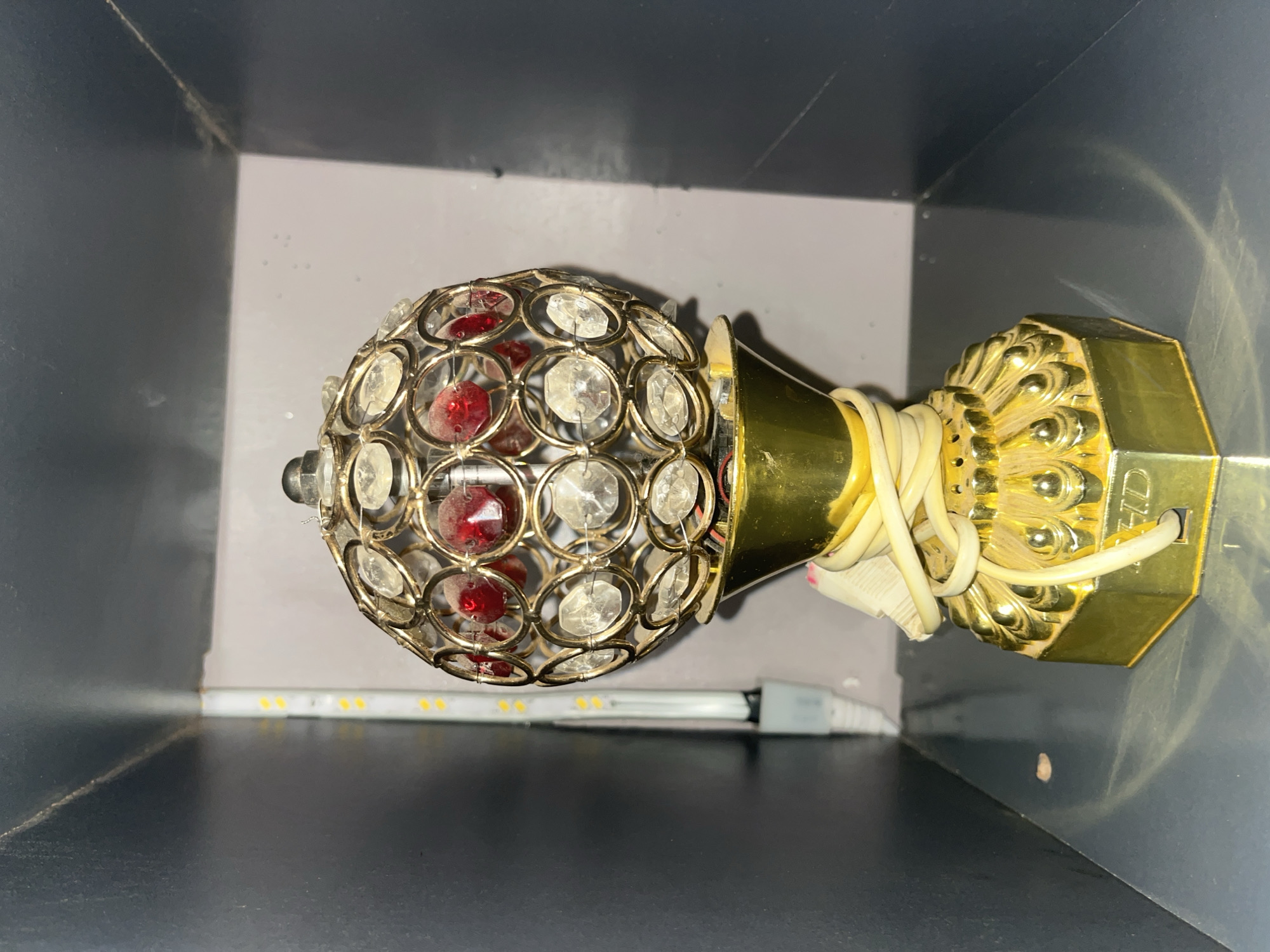 The image shows a decorative lamp with a gold-colored base and a spherical top adorned with clear and red crystals. The lamp is placed on a reflective surface inside a dark, compartmentalized shelf or cabinet. A white power cord is wrapped around the base of the lamp. In the background, on the left side, a strip of LED lights is visible, partially obscured by the shelf's structure. The overall impression is that of an indoor setting, likely a shelf in a home or storage area. The specific location in Jos, Nigeria, cannot be determined from the visual information provided.