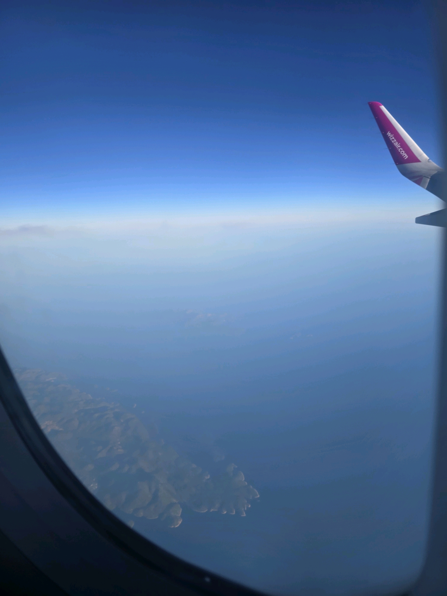 An aerial view from an airplane window reveals the island of Hvar, Croatia, appearing as a rugged, mountainous landmass partially submerged in the deep blue sea. The coastline is irregular, with bays and peninsulas dotting the perimeter. The terrain is characterized by a lack of distinct human settlements visible from this altitude, presenting a primarily natural landscape. The sky above is a gradient of deep blue, transitioning to a lighter hue near the horizon where it meets a thin band of hazy clouds. On the right side of the frame, the pink and white wingtip of the airplane is visible, bearing the text "wizzair.com". The window frame itself creates a dark, curved border around the view.