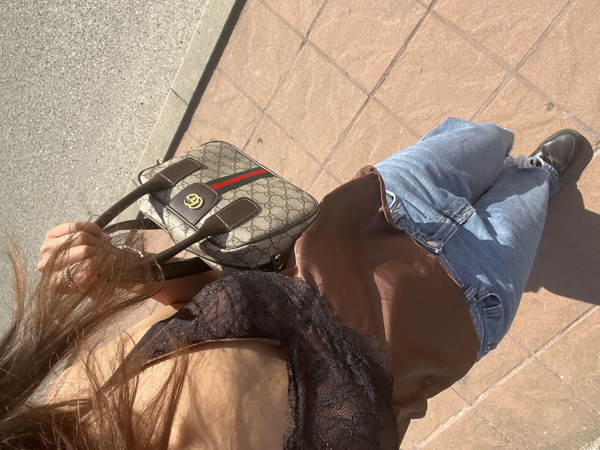A person is captured from a high-angle, overhead shot, outdoors on a sunny day. They are seated on a paved area made of tan square tiles, with a textured grey surface bordering it. The person is wearing blue denim jeans with ripped details and a brown top. A black lace garment is visible over the brown top, extending to the chest area. The person is holding a designer handbag, identifiable by its monogram pattern and the green and red stripe detail. A gold-toned "GG" logo is prominently displayed on the bag. Their long, brown hair cascades over their shoulder and down their arm. A shadow is cast on the tiled ground, suggesting a clear, sunny atmosphere. The individual's right foot is visible, wearing a black shoe with white laces. The scene appears casual and relaxed. No visible text is present in the image. The location context is Zlatograd, Bulgaria.