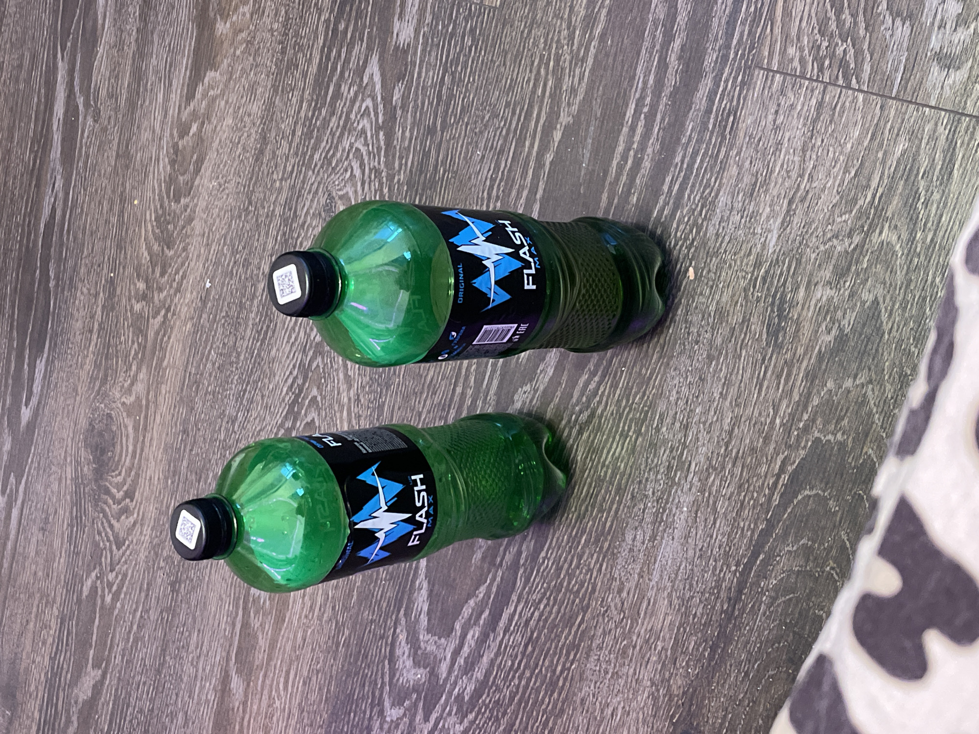 Two green plastic bottles of "FLASH MAX ORIGINAL" energy drink are placed on a wooden floor. Each bottle has a black cap with a small QR code sticker on top. The bottles are partially filled with a dark green liquid. The floor has a visible wood grain pattern and appears to be indoors. There are no people or animals present in the image. The scene is static and does not depict any action. The lighting suggests an indoor environment, possibly a home or store.