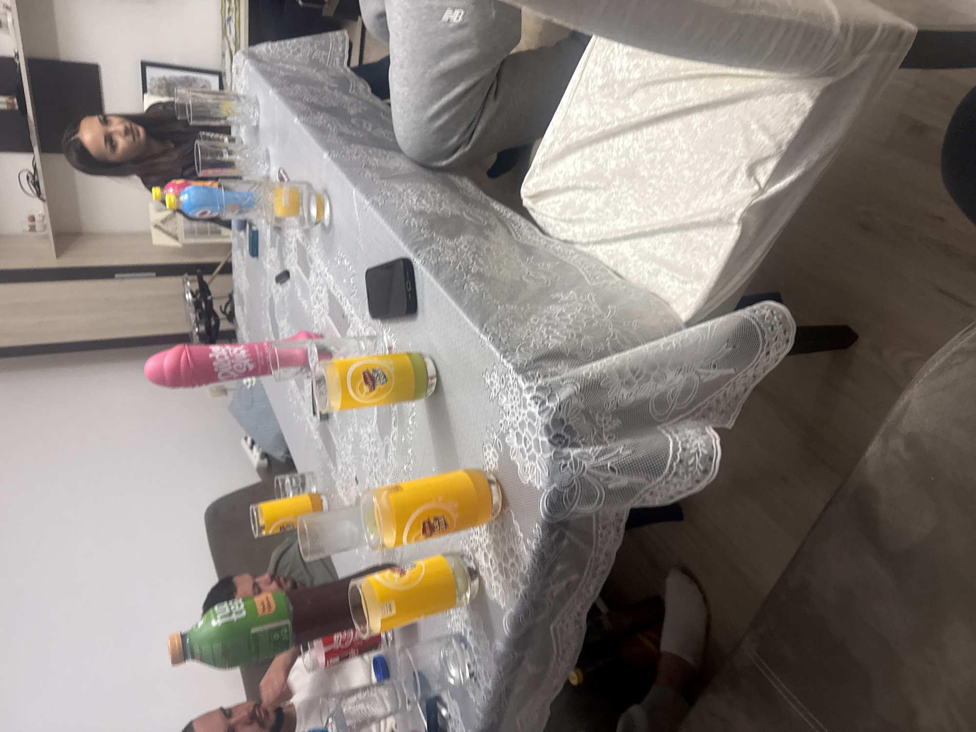 The image shows a group of people seated around a table covered with a white lace tablecloth. The table is cluttered with glasses and bottles of various beverages, including orange juice, soda, and mineral water. A black mobile phone sits on the table. People are seated around the table, and one is wearing grey sweatpants with a New Balance logo. The location could be Graf Ignatievo, Bulgaria, but there is no evidence in the image to confirm this.