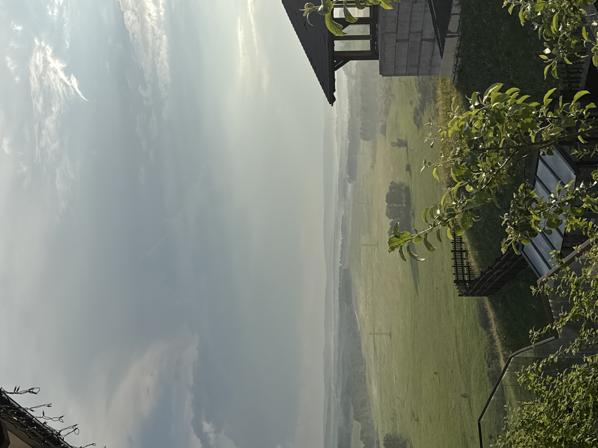 This image depicts a scenic landscape with a hint of human habitation in the foreground and middle ground, under a vast, cloudy sky. The location is identified as Shamoni, Russia.
In the foreground, to the right, a portion of a building with a dark, angled roof and light-colored brick walls is visible. The roof has some details of exposed beams. In front of this structure, a lush green leafy branch extends into the frame, obscuring part of the building. Below and to the left of this branch, more greenery and the tops of glass-roofed structures are visible, suggesting a garden or greenhouse area. A dark wooden fence runs along a sloping grassy hill, separating the immediate foreground from the wider landscape.
The middle ground reveals a sprawling green valley or meadow, dotted with trees and possibly some scattered structures or power lines. A light mist or fog hangs low over the landscape, softening the distant hills and creating a hazy atmosphere. The sun appears to be either rising or setting, as indicated by the soft, diffused light and the atmospheric haze.
The background consists of layers of distant hills fading into the haze, with a dramatic sky above. The sky is a mix of light and dark clouds, with patches of brightness suggesting sunlight breaking through. There are also some string lights hanging from the overhang of the building in the top left corner of the image.
The overall impression is one of a tranquil, rural setting, possibly a residential area with a view of the natural landscape. There are no people or animals visible in the image. The scene is captured in natural daylight, with a slightly muted color palette due to the atmospheric conditions.