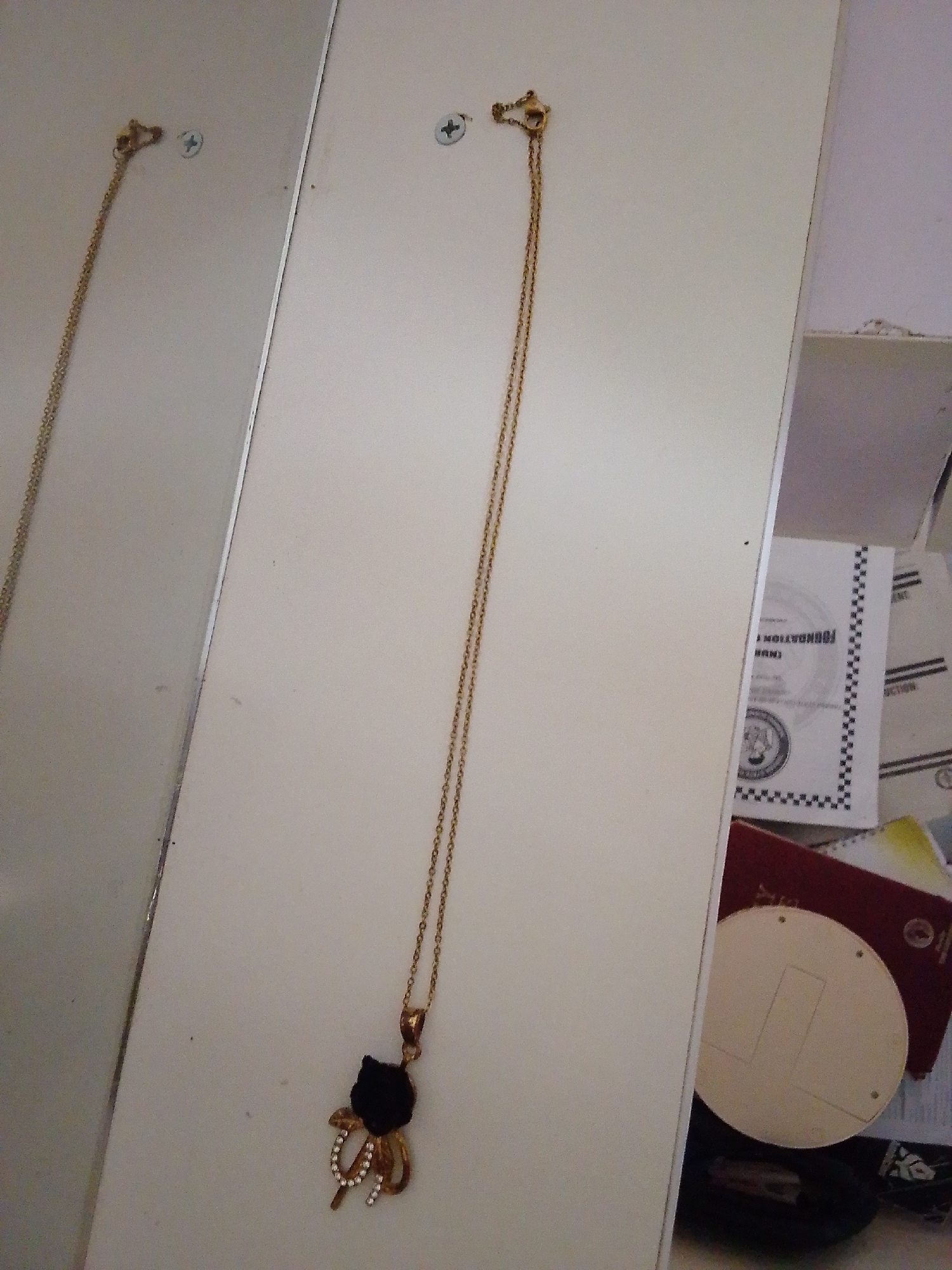 A gold necklace with a black cat pendant adorned with small white stones hangs from a hook on a white wall. The necklace is positioned vertically, extending from the top of the frame downwards. To the left, a reflection of the necklace and a portion of the wall is visible in what appears to be a mirrored surface. The wall has two visible screws. In the background on the right, stacked items, including a red folder or book, a white booklet with text and a checkered border, and other papers, are partially visible. The scene is illuminated with natural light, casting soft shadows. The location is identified as Kuson, Nigeria.