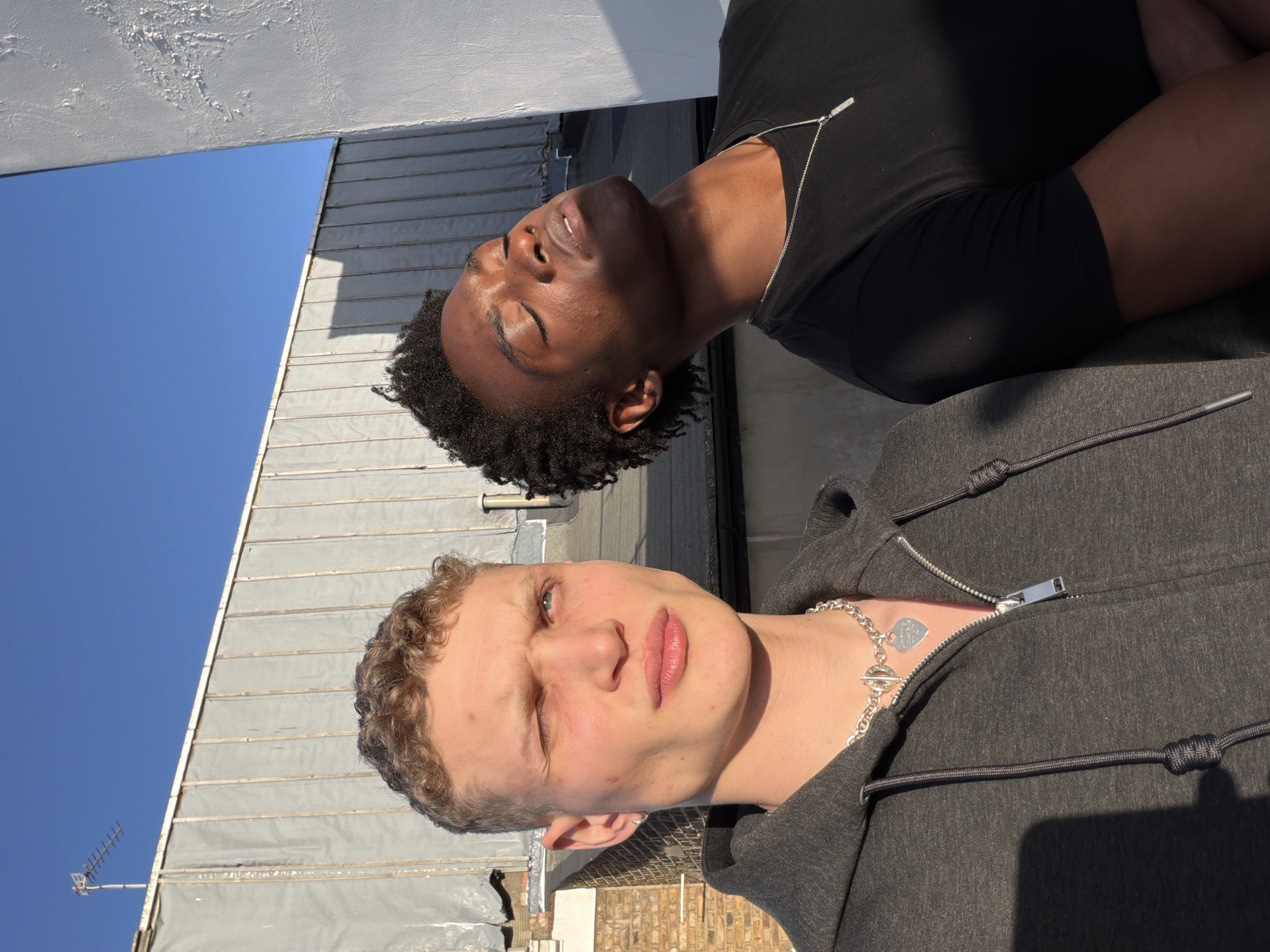 Two young men are posing for a selfie outdoors on a sunny day. The man in the foreground has fair skin, light-colored curly hair, and is wearing a gray zip-up hoodie and a silver chain necklace with a heart pendant. He is squinting slightly, and his lips are closed. The man in the background has dark skin, short dark curly hair, and is wearing a black t-shirt and a silver chain necklace. His eyes are closed, and he seems to be looking up. They appear to be on a rooftop with a corrugated metal surface, possibly in London, United Kingdom, as suggested by the presence of brick buildings and an antenna in the background. The sky is clear and blue.