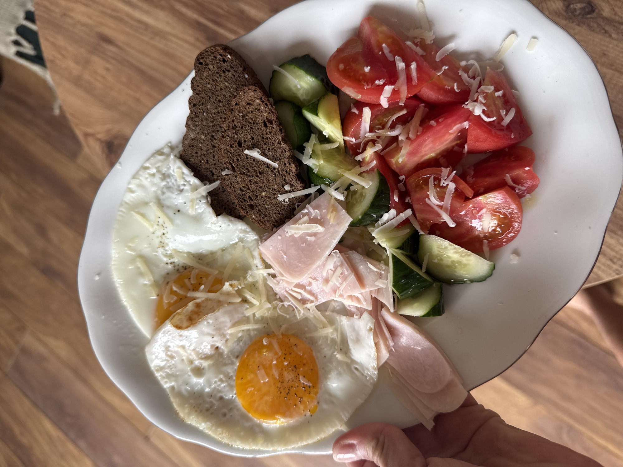 A plate of food is displayed on a wooden surface, possibly a table. The food consists of two fried eggs with yolks, a slice of dark bread, slices of ham, and a salad with cucumbers and tomatoes. The salad and eggs are topped with grated cheese. A hand is holding the plate. The location appears to be indoors.