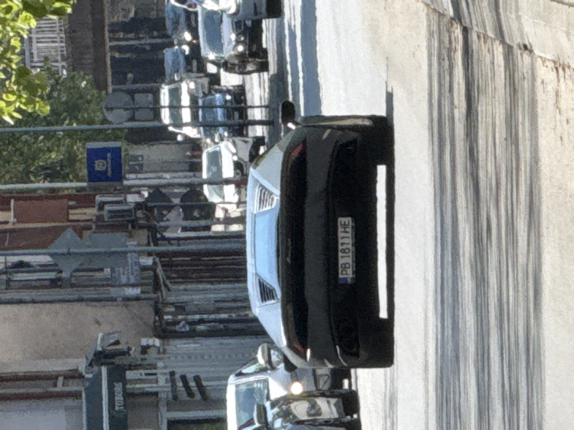 The image shows a street scene in Plovdiv, Bulgaria, featuring a black car with license plate PB 1811 HE. The car is parked on the right side of the road, next to a sidewalk with vertical shadows cast from an adjacent building. Other vehicles, including a white van and several cars, are visible further down the street, indicating moderate traffic. A Husqvarna sign is visible in the background to the left of the image. The presence of trees suggests an urban environment. The image appears to have been taken during daylight hours.