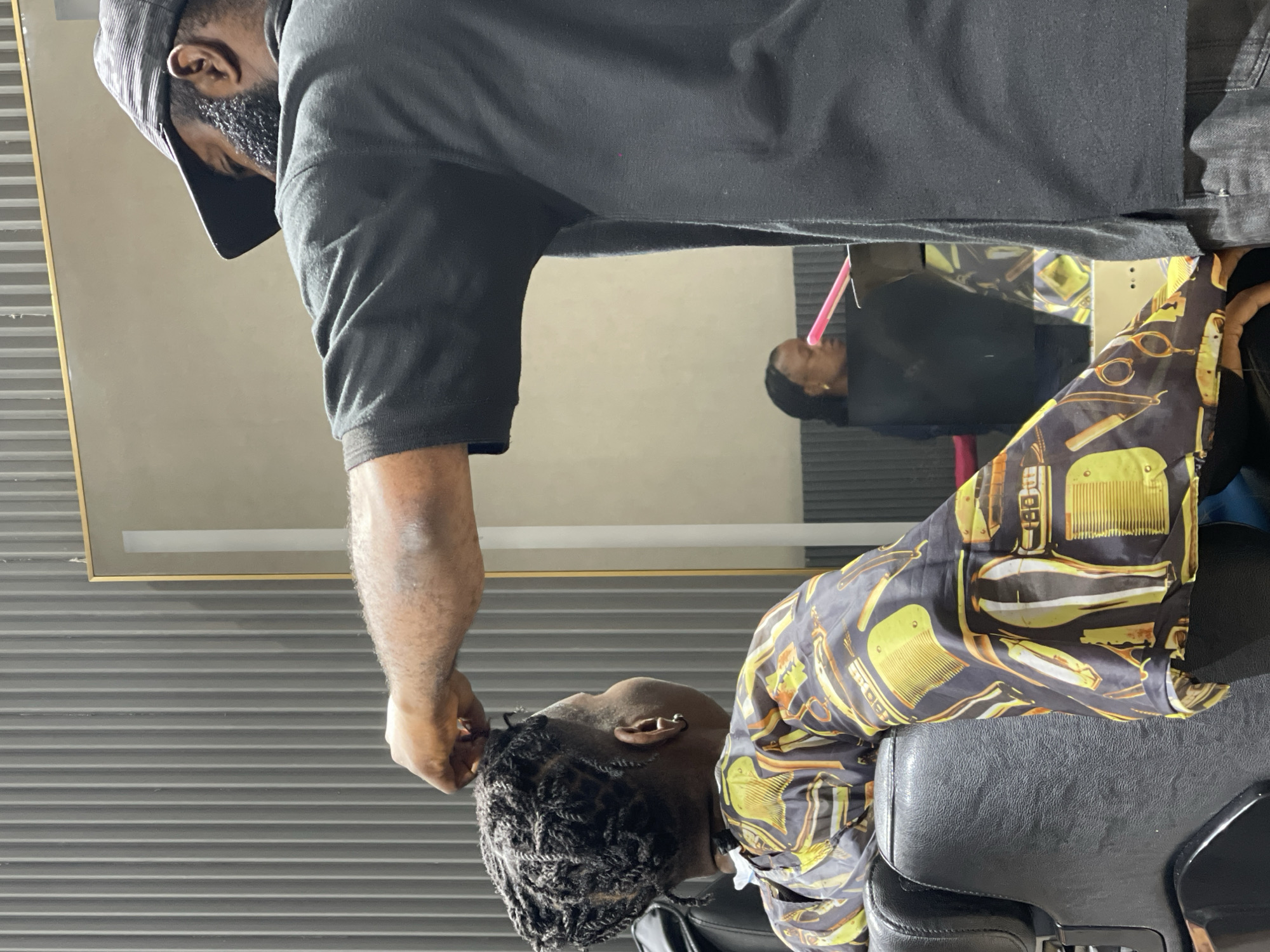 In a barbershop in Guzape, Nigeria, a barber is styling a client's hair. The barber, wearing a black baseball cap and a black t-shirt, is focused on the client's braided hair. The client is seated, wearing a patterned cape with images of barbershop tools like combs and scissors. The background shows a mirror reflecting another person, possibly another client, and a wall with horizontal grey lines. The overall atmosphere suggests a typical day at a barbershop.