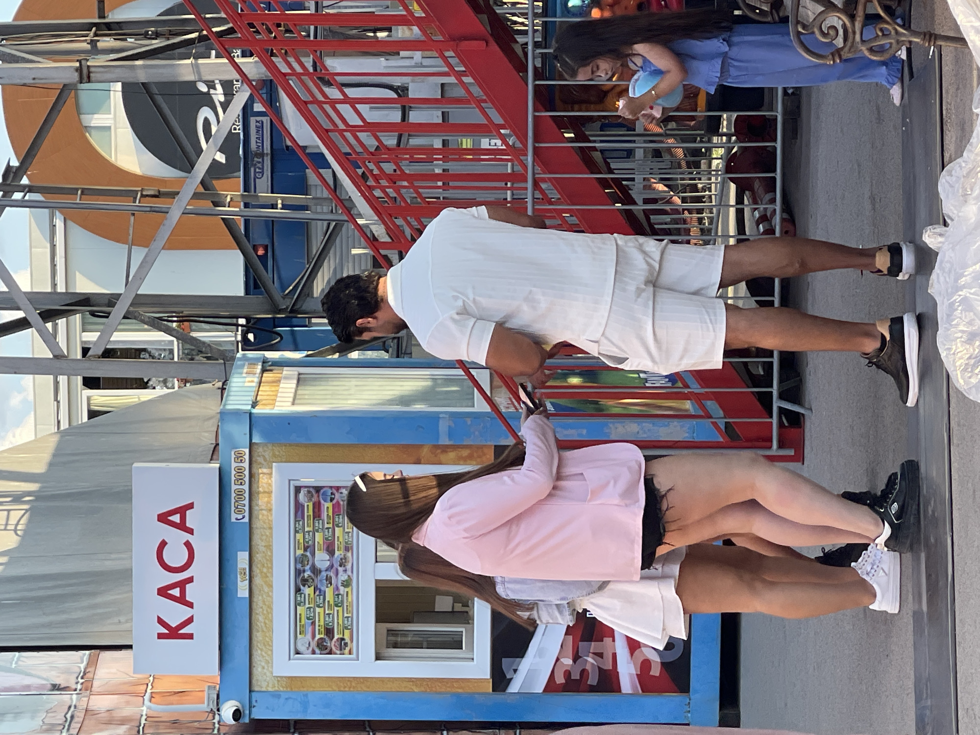 The image shows a street scene in the city of Varna, Bulgaria, possibly near an amusement park or fairground. In the foreground, a man in a white short-sleeved collared shirt and white shorts stands facing away from the camera. To his left, two women are partially visible. One woman, wearing a pink jacket and dark shorts, holds a smartphone. The other woman, behind her, wears a light blue denim jacket and a white skirt. Further to the right and in the background, a young girl in a blue outfit is holding a blue object.
Behind the people, there is a blue and yellow booth with a sign that reads "KACA" in red letters. Below the sign is a phone number. A window on the booth displays pictures and prices. To the right of the booth, a red staircase structure is visible, with metal scaffolding. In the far background, a building with an orange arch is present. The ground appears to be paved.