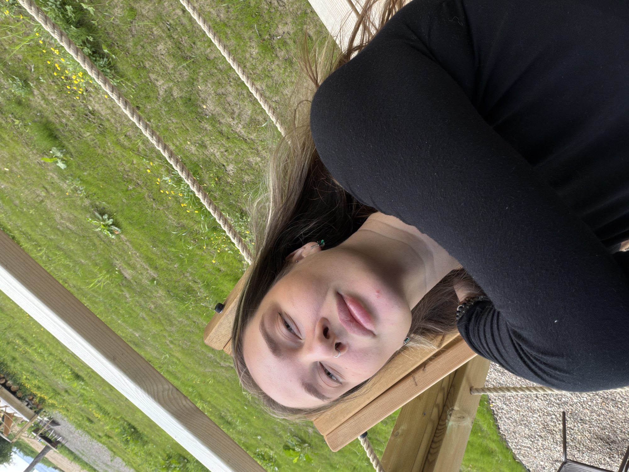 A young woman with long, light brown hair is lying on a wooden bench, her body angled towards the camera. She is wearing a black long-sleeved shirt. Her head is tilted back, and she is looking at the camera with her eyes partially closed. She has a nose ring and is wearing small green earrings. Her left arm is bent, resting across her chest. The bench appears to be part of an outdoor structure, possibly a swing or a hammock, made of light-colored wood and supported by ropes. The background is a grassy area with scattered yellow flowers and some patches of bare ground. In the distance, there are more wooden structures and a body of water is visible under a bright sky. The scene suggests a relaxed outdoor setting. The location is identified as Solov'evo, Russia.