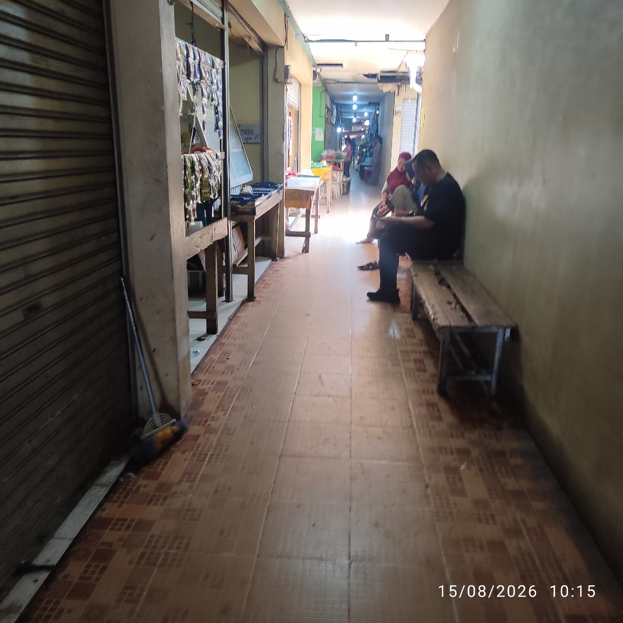 The image shows a narrow, indoor walkway or hallway with tiled flooring. On the left, a metal roller shutter door is visible, partially covering a storefront display. The storefront features shelves with various items, possibly snacks or small goods, in foil packaging. To the right of the walkway, there is a light-colored wall and a rustic wooden bench. A few people are seated on the bench, including a person wearing a black t-shirt and dark pants, who appears to be looking down at a phone or tablet. Another person, wearing a red headscarf, is sitting next to them. The walkway extends into the distance, where more stalls and individuals are faintly visible, illuminated by overhead lighting. The floor is covered with square brown and beige tiles, some of which appear worn or stained. A broom with a yellow and blue brush is leaning against the wall near the shutter door. The overall atmosphere suggests a market or a commercial area with pedestrian traffic. The timestamp "15/08/2026 10:15" is visible in the lower right corner, indicating the date and time the photo was taken. The location is identified as Pembaharuan, Indonesia.
