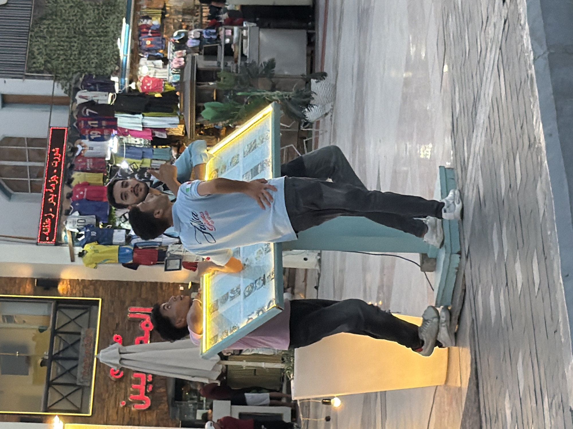 In Sharm El Sheikh, Egypt, three young men are gathered around a lighted menu board. One man, wearing a pink shirt and dark pants, leans over the board, looking up and to his left with an animated expression. He is standing on what appears to be a wooden plank, which is itself on top of a lower platform. Another man, in a light blue shirt with an Italian flag detail on the sleeve, stands facing right, his left hand on his hip and his right hand raised as if gesturing. He is looking towards the first man. A third man, wearing a darker shirt, stands behind the menu board, his face turned towards the camera, and he is giving a thumbs-up gesture. The menu board displays images of food items. The scene appears to be an outdoor setting, possibly a restaurant or cafe, with shops selling clothing visible in the background. The shops are stocked with t-shirts and other apparel, some featuring numbers. The lighting suggests it is either evening or a shaded area.