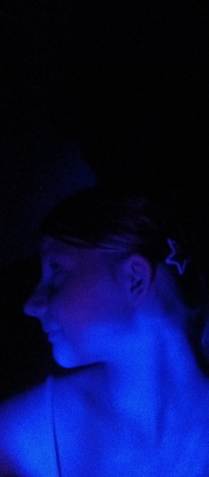 A person is seen in profile, illuminated by a blue light source. A star-shaped hair clip is visible in their hair. The environment is dark, obscuring any specific details about the location, which is thus listed as Unknown, Unknown. The individual's face is largely in shadow due to the lighting conditions.