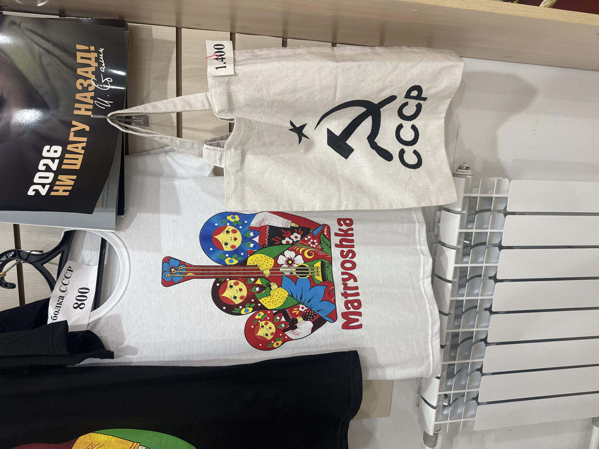 In an indoor setting, likely a shop or market stall in Sochi, Russia, several items are displayed. To the left, a black t-shirt hangs from a hanger. To its right, a white t-shirt is displayed, featuring a colorful graphic of Matryoshka dolls with a guitar and the text "Matryoshka" below. Above the white t-shirt, a price tag reads "800" and some Cyrillic text that translates to "T-shirt USSR". To the right of the white t-shirt, a light-colored canvas tote bag with the hammer and sickle symbol and "CCCP" is hanging. A price tag reading "1.400" is attached to the bag. In the background, a poster with the text "2026 НИ ШАГУ НАЗАД!" (2026 NOT ONE STEP BACK!) and a signature is visible. A white radiator is situated at the bottom of the image. The background appears to be a light wood-paneled wall.