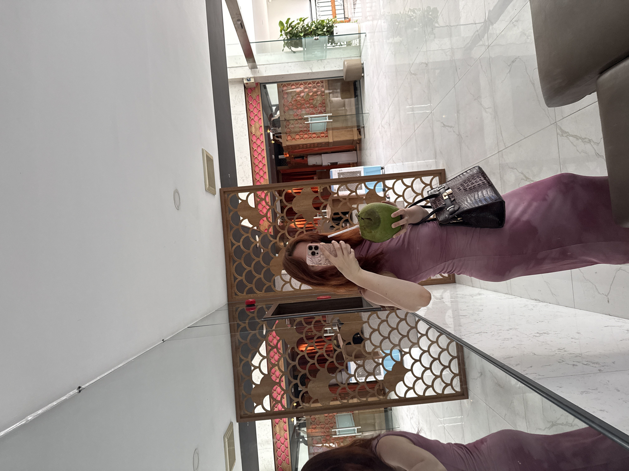 A red-haired woman is standing indoors, taking a selfie with a smartphone. She holds a green coconut in her left hand. The woman is wearing a purple dress and has a black purse resting against her hip. The background features an ornate, geometric wooden screen, a marble floor, and partial view of an indoor atrium with a glass railing and plants. The photograph was taken inside a reflective surface, as indicated by the mirrored image of the woman and her surroundings. The location is Bac Nha Trang Ward, Vietnam.