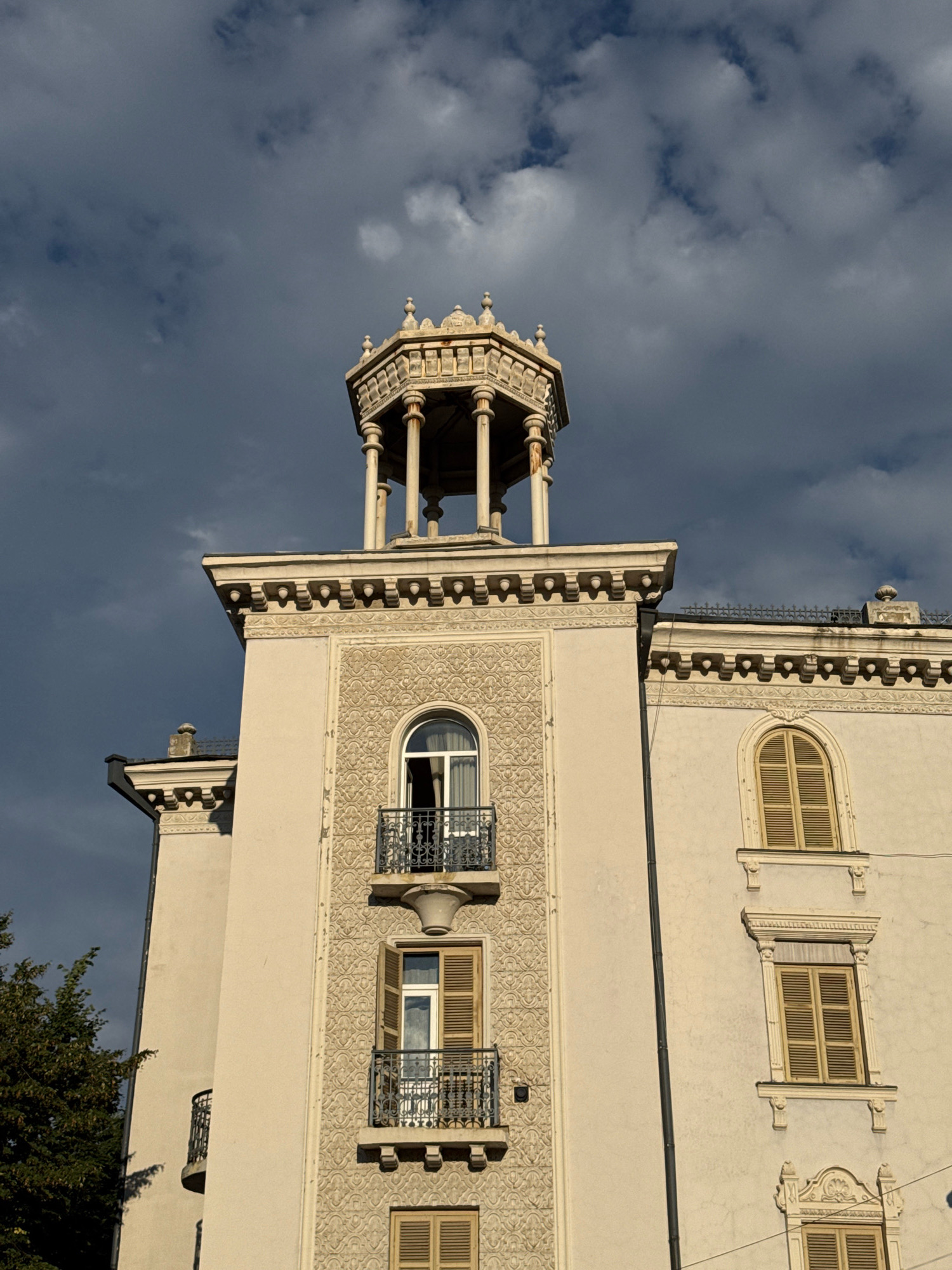 The image captures an upward view of the upper facade of a light-colored building, possibly an apartment or hotel, with a distinctive architectural style that includes ornate detailing and a turret-like structure at the very top. The building's facade is decorated with intricate relief patterns. Several windows are visible, some with balconies featuring black metal railings. Some windows are open, revealing curtains, while others are covered with light-colored wooden shutters. The sky above is dark and cloudy, with patches of lighter sky and sunlight filtering through, suggesting a dynamic weather condition. To the left of the building, the dark green foliage of a tree is partially visible. The scene appears to be situated in the city of Telavi, Georgia. There are no people or animals visible in the image.
