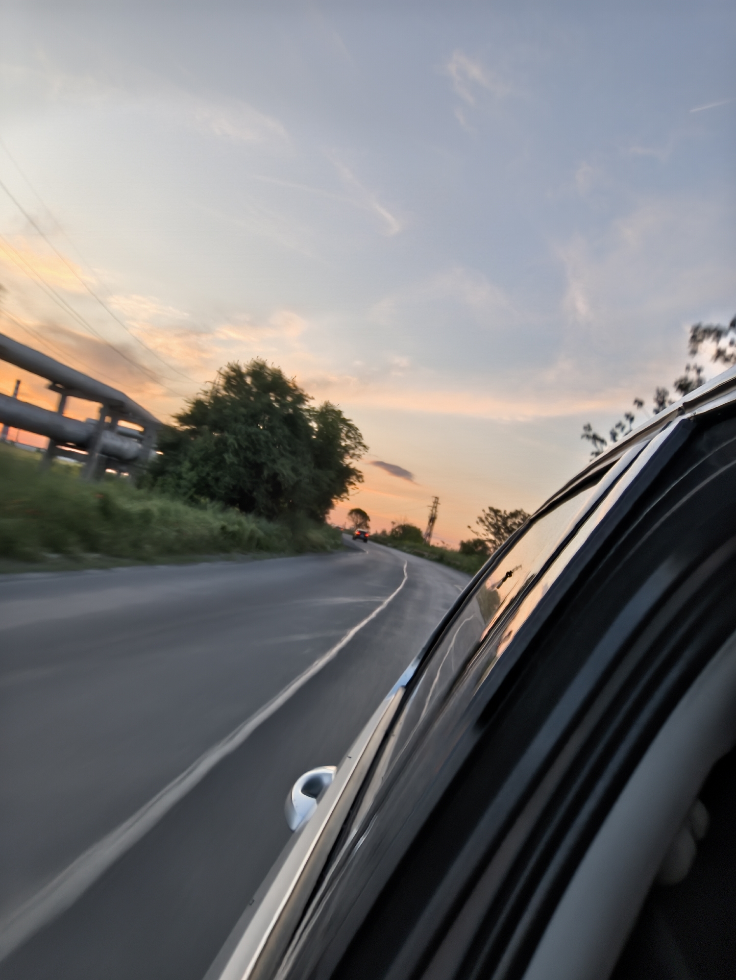 The image depicts a view from inside a moving car on a road at dusk or dawn. The sky is painted with shades of orange and blue, suggesting either sunrise or sunset. A road with a white dividing line stretches into the distance. On the left side of the road, there is a patch of greenery with trees and what appears to be an industrial pipeline structure. In the distance, another vehicle is visible on the road ahead. The foreground is dominated by the interior of the car, showing the window and frame. The image does not definitively place the location in Burgas, Bulgaria, so the city is not verifiable from the image alone.