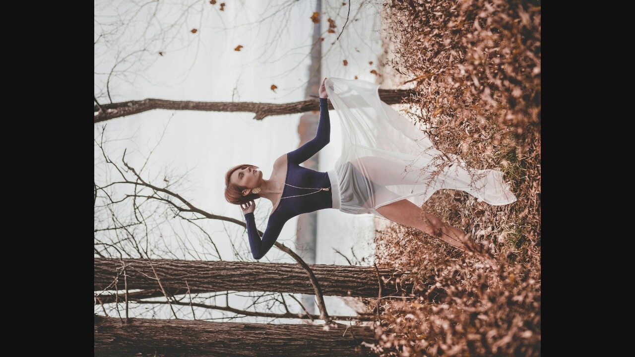 A full-shot, medium-toned photograph shows a woman with short, reddish-brown hair posing outdoors in a wooded area near a body of water. She is wearing a dark blue, off-the-shoulder long-sleeved shirt and a sheer white skirt with a thigh-high slit. A long, thin necklace with a gold pendant hangs from her neck. Her left arm is bent at the elbow with her hand behind her head, and her right arm is extended, with her hand near her skirt. Her left leg is bent at the knee, and her right leg is extended slightly forward. She is looking to her right. The background consists of bare trees, dry brush and leaves on the ground, and a calm body of water with a muted, overcast sky beyond. The overall lighting is soft and diffused, suggesting an outdoor setting with overcast weather. The scene is devoid of any urban elements, indicating it is likely in a natural or rural environment.