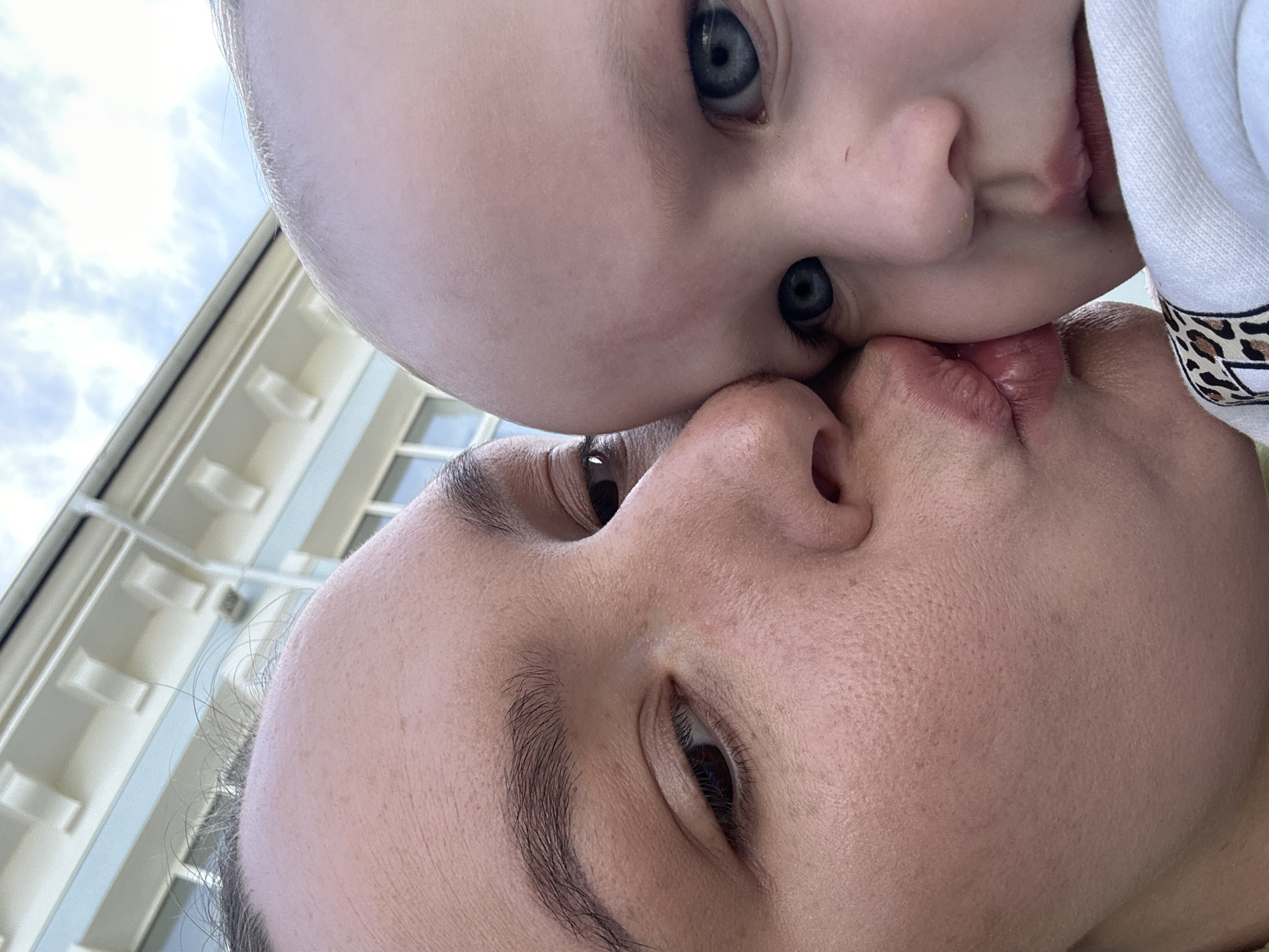 A close-up, low-angle shot captures the faces of a young child and an adult. The child, positioned above, has fair skin, blue eyes, and is wearing a white garment with an animal print trim. The adult below has brown eyes and dark hair. Their faces are close, almost touching, with their lips meeting. The background reveals the exterior of a building with a multi-story facade, possibly located in Chaikovsky, Russia. The sky, slightly visible, appears cloudy.