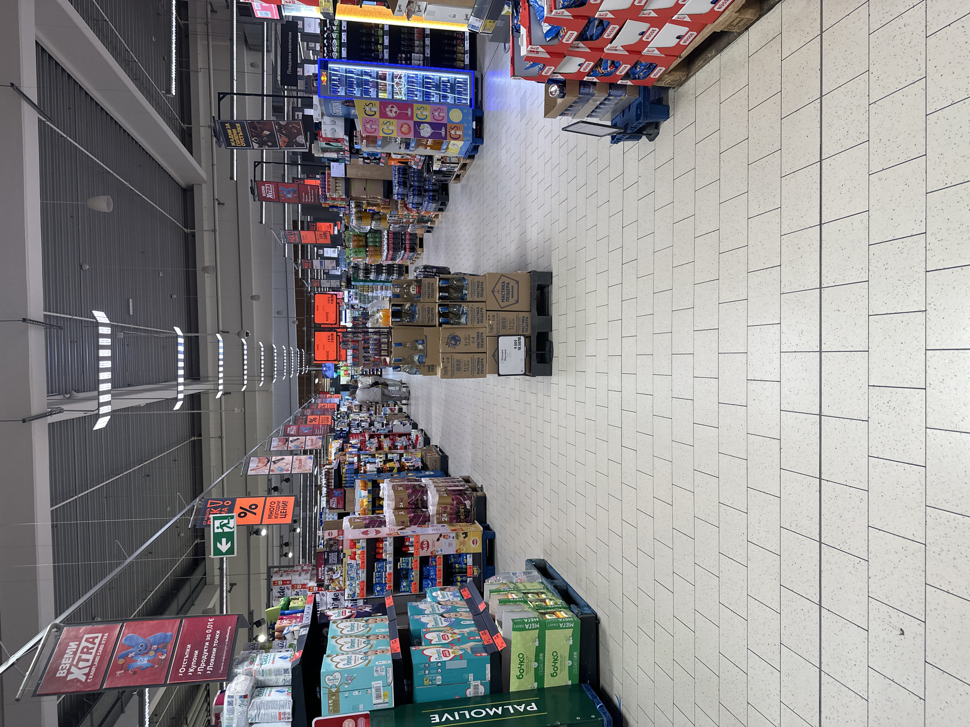 The image displays a wide-angle view of a brightly lit, long aisle inside a supermarket, likely a Kaufland store given the visible signage. The perspective looks down the aisle from foreground to background.
On the left side, shelves are stocked with various products, including a prominent, brightly lit Red Bull cooler and other beverages. Further along, more goods are stacked on pallets. Numerous hanging signs are suspended above the aisle. On the right side, products like Pampers diapers, "БОЧКО" (Bochko) brand items (some marked "МЕГА ПАКЕТ" - MEGA PACK), and "PALMOLIVE" personal care products are displayed in bulk on pallets. Near the background, a blue cart loaded with red boxes labeled "NÜSSE" is visible.
The floor is made of light-colored, speckled rectangular tiles. The ceiling features exposed corrugated metal decking and linear fluorescent light fixtures, providing uniform illumination. No people are visible, and the aisle appears well-organized and stocked.
Visible text on signs and product packaging includes:
* "ВЗЕМИ XTRA С KAUFLAND CARD XTRA" (GET XTRA WITH KAUFLAND CARD XTRA)
* "ЖАДНИ ЗА ГОЛЕМИ отстъпки" (THIRSTY FOR BIG discounts)
* "Много Изгодни ЦЕНИ!" (Very Favorable PRICES!)
* "Плодови напитки" (Fruit drinks)
* An exit sign (green with white figure and arrow).
* A sign listing: ">Отстъпки >Купони >Продукти за 0,01€ >Лоялни точки" (>Discounts >Coupons >Products for 0.01€ >Loyalty points).
* Product labels show "PALMOLIVE", "БОЧКО", "Pampers", "Red Bull", and "МАСТИКА ПЕЩЕРА" (Mastica Peschtera) boxes, some indicating "6x1L", "9.50€", and "18.58ЛВ." (18.58 Bulgarian Lev).
The prevalence of Bulgarian text on signs and products, such as "Мастика Пещера" and "Бочко", strongly suggests the location is in Bulgaria.