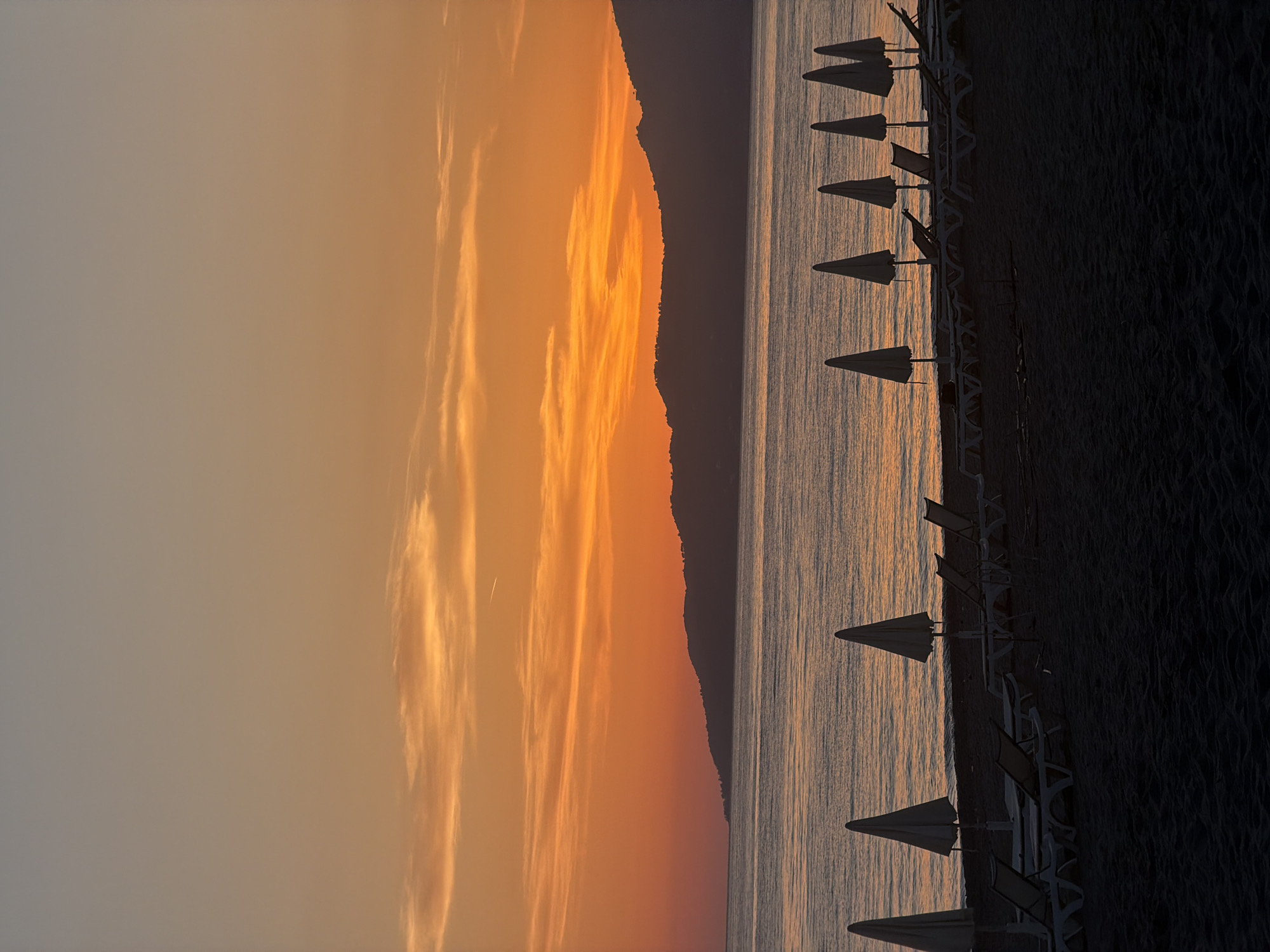 This image depicts a sunrise scene at Ascea Marina in Ascea, Italy. The sky displays an orange gradient with striated clouds. A mountain silhouette separates the sky from the sea, which reflects the orange sky. A row of white sun umbrellas lines the beach. The beach itself is dark, possibly sand. No people are visible.