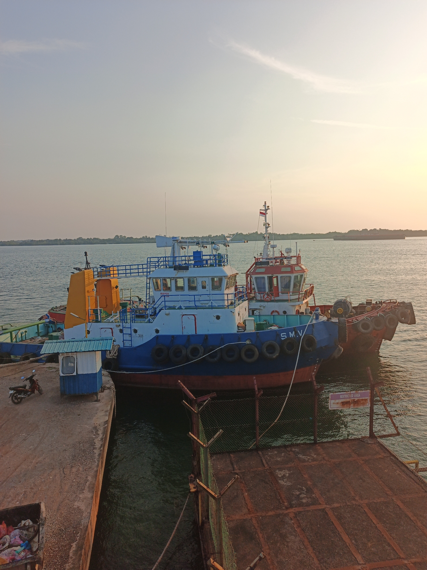 The image depicts a waterfront scene at a port, likely in Batam, Indonesia, given the presence of tugboats and the general industrial maritime atmosphere. The time of day appears to be late afternoon or early evening, with a soft, warm light casting long shadows.
In the foreground, a concrete pier juts out into the water. On the pier, a small, weathered blue and white booth stands near the edge, with a red motorcycle parked beside it. To the left of the pier, a dark metal dumpster is filled with various refuse, including plastic bags and debris.
Dominating the midground are two tugboats moored alongside the pier. The tugboat closer to the viewer is painted blue and red, with "S.M.V." clearly visible on its side. It is adorned with numerous black tires along its hull for protection. The second tugboat, partially obscured behind the first, is also blue and white, with a distinctive yellow structure at its rear. Both vessels appear operational, though their exact purpose at this moment is unclear.
The water surrounding the pier and boats is a murky green, with gentle ripples suggesting a calm day. In the distance, the coastline is visible, lined with trees and vegetation. Further out on the water, a large barge or cargo ship can be seen sailing on the horizon, silhouetted against the pale, hazy sky. A tall antenna or mast extends upwards from one of the tugboats.
A rusted metal fence with a green mesh runs along the edge of the pier in the right side of the image, leading to a metal platform. A small sign with a purple border and white text, reading "RESTRICTED AREA DO NOT ENTER", is attached to the fence. The overall impression is one of a working port, possibly at the end of a day's operations, with the gentle light creating a serene, yet functional, atmosphere.