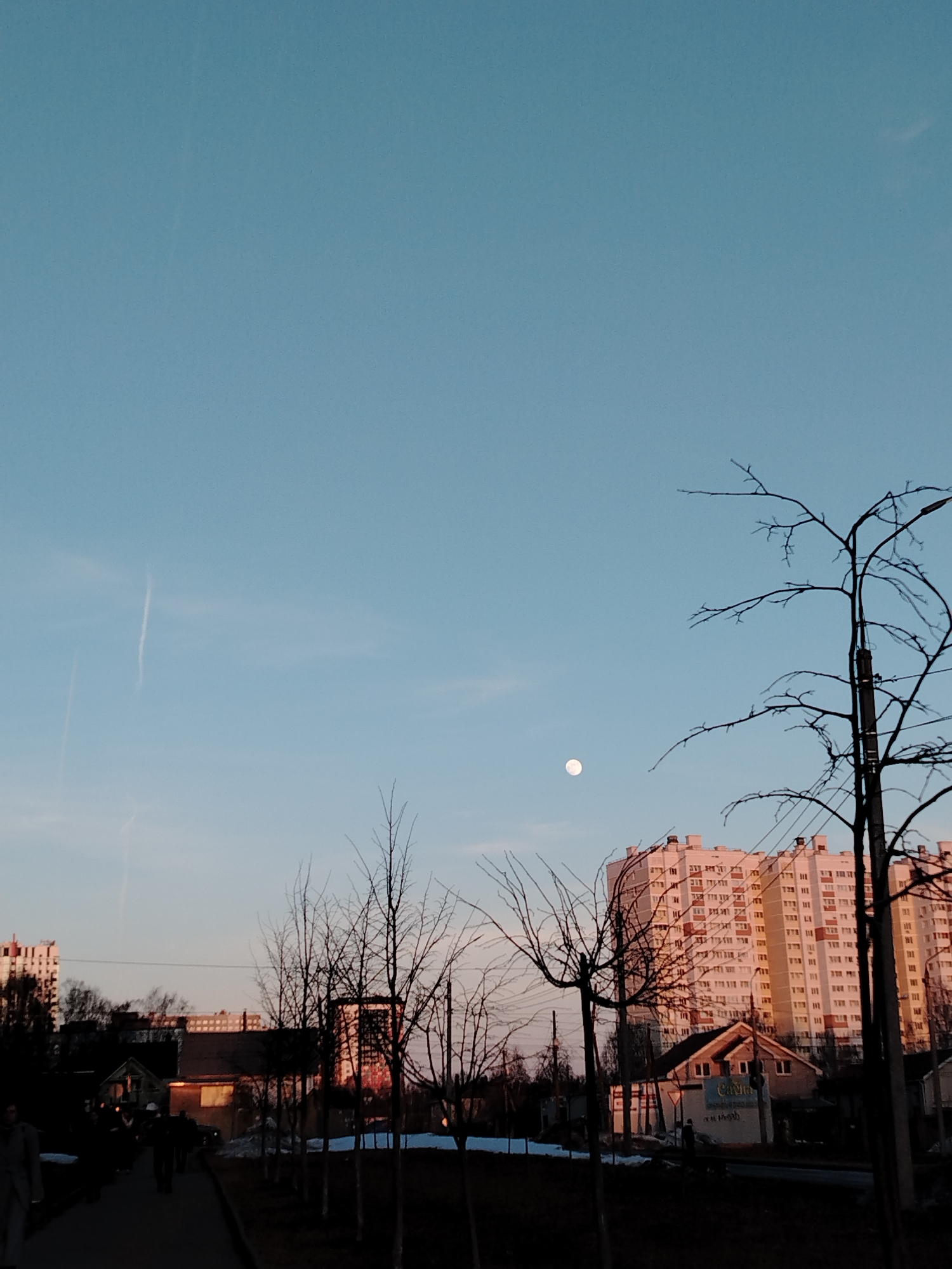 The image depicts an urban scene in Izhevsk, Russia at dusk. A light blue sky with faint contrails serves as a backdrop for a mixed landscape of residential buildings, bare trees, and streetlights. In the foreground, dark silhouettes of people walk along a sidewalk. Behind them are smaller buildings, some with visible signage. A large, multi-story apartment building, illuminated by the setting sun, dominates the middle ground. The moon is visible in the sky between the buildings and branches. The scene suggests a transitional time of year, possibly late autumn or early spring, with no foliage on the trees. Patches of snow remain on the ground. The atmosphere is somewhat muted, with the setting sun casting long shadows.