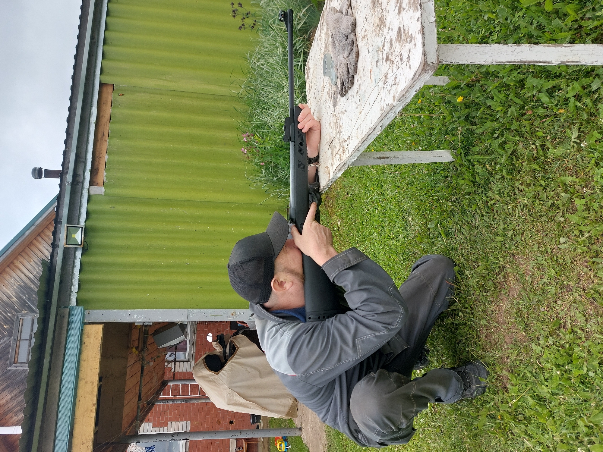 The image captures an outdoor scene in Большой Букор, Russia, under an overcast sky. A single adult male, approximately 30-40 years old, is depicted in a low crouch, with his left knee on the grassy ground and his right foot flat. He is oriented towards the right, actively aiming a black rifle with open sights. The rifle's forend is supported by his left hand, resting on a weathered, rectangular wooden table with peeling white paint. The man wears a black mesh baseball cap, a two-tone grey work uniform (light grey upper section, darker grey lower section and sleeves), dark grey cargo pants, and black athletic shoes. His right eye is aligned with the rifle's sights. On the right side of the table, a single grey fabric work glove and a small, metallic, cylindrical container are visible.
In the midground, behind the individual and the table, a corrugated green metal structure, likely a shed or wall, forms the primary backdrop. A horizontal wooden beam is affixed across its upper section. An LED floodlight is mounted on the green structure to the far right. Further behind, to the left, a portion of a red brick building with a visible window is present, and a white ladder leans against an adjacent structure. A light-colored tarp or cover obscures an object immediately behind the individual and to the left. The upper section of a wooden-sided building with a pitched roof and a small attic window is visible above the green structure. A dark-colored metal chimney pipe with a cap extends from a roofline in the background. The ground consists of green grass with interspersed weeds, including dandelions and darker flowering plants, with some areas of exposed soil in the foreground.