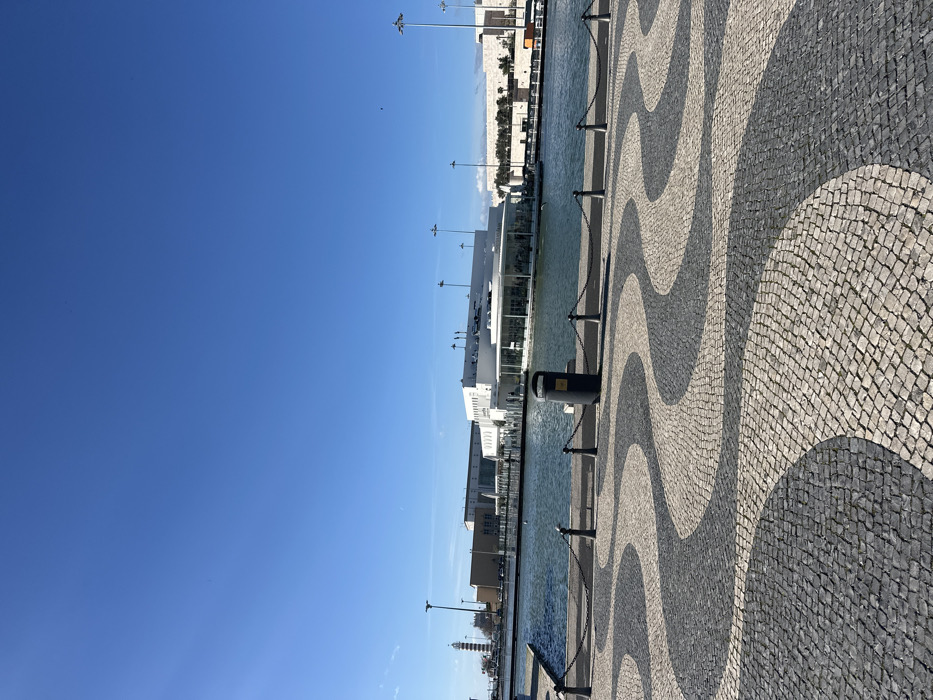 The image displays a waterfront scene in Parque das Nações, Lisbon, Portugal, under a clear, bright blue sky. The foreground is dominated by a Portuguese mosaic pavement (Calçada Portuguesa) featuring an intricate wavy pattern composed of alternating dark grey and light grey/white cobblestones. A dark metal barrier, consisting of short posts connected by chains, runs horizontally across the lower midground, separating the paved area from a calm body of water. Positioned behind this barrier is a dark grey cylindrical public waste bin with a yellow label.
Across the water, a prominent modern white building with a distinctive curved glass facade, identifiable as the Lisbon Oceanarium, forms the central visual element of the midground. To the left of this building, further along the waterfront, a red and white striped cylindrical lighthouse with a conical top is clearly visible. Various dark-colored lampposts are scattered across the midground, both near the water's edge and behind the buildings. The sky above is devoid of clouds, exhibiting a uniform deep blue color, with a slight sun glare visible in the upper left quadrant. No discernible individuals or specific actions are clearly depicted in the scene.