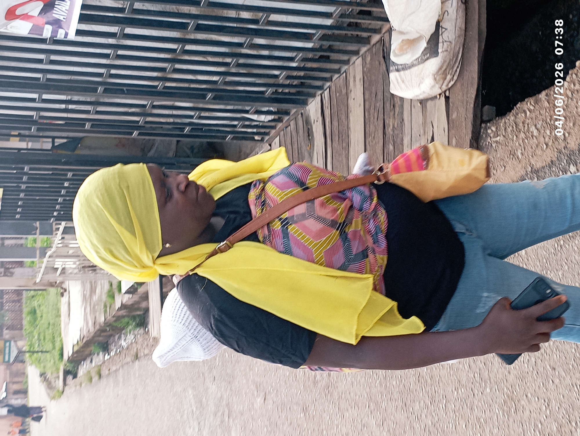 This image, taken in Jos, Nigeria, captures a woman standing outdoors on an unpaved road or path. The timestamp "04/06/2026 07:38" indicates it's early morning.
The central subject is a woman, seen from the waist up, dressed in a bright yellow headscarf that covers her hair and neck. She wears a black long-sleeved top, layered with a colorful outer garment featuring pink, yellow, and black geometric patterns. Her lower body is clad in light blue jeans, and she carries a yellow-orange handbag with a brown strap across her body. In her left hand, she holds a dark-colored smartphone. Her face is mostly visible, with a calm, neutral expression as she looks slightly away from the camera.
The background features a metal slatted structure, possibly a gate or storefront, directly behind the woman. Beyond this, a rough wooden platform or wall is visible. To the right, a white sack labeled "SALT" is resting on the wooden structure. The ground beneath her is a dusty, unpaved surface. In the distance, on the left side of the frame, there are faint hints of greenery and some indistinct structures, suggesting a street or pathway in a semi-urban setting. A partial sign with the word "AVAILABLE" can be seen in the upper left corner of the background. The lighting is soft, consistent with an early morning scene.