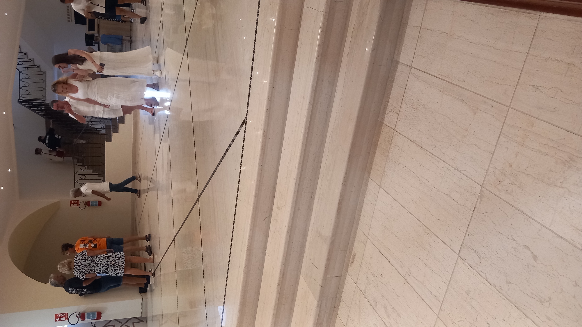 The image shows a wide shot of an indoor area, likely a hotel lobby or a public building, with people milling about. The flooring consists of large, light-colored marble tiles, with darker inlays forming geometric patterns. In the foreground, there are several wide, shallow marble steps leading up from the tiled floor.
To the left, a group of four people are conversing. A man with graying hair, wearing a black t-shirt and shorts, stands with his back to the camera. Next to him, a woman in a black and white patterned dress and sandals is facing the camera. To her right, a young person in an orange t-shirt and shorts is also facing the camera. Beside them, a man in a white t-shirt and dark shorts is partially visible.
Further back, on the right side of the image, a group of three women and two men are walking together. The women are all dressed in white. One is wearing a white dress, another a white tank top and a long white skirt, and the third a white dress. The men are wearing white shirts and shorts.
In the background, a staircase with a dark metal railing is visible. There are other people on the staircase, including a person in a black hoodie. Fire extinguishers are mounted on the wall in two locations. The overall environment appears to be well-lit and spacious, with an arched ceiling. The scene is likely taking place in Kos, Greece, possibly in a hotel or resort.