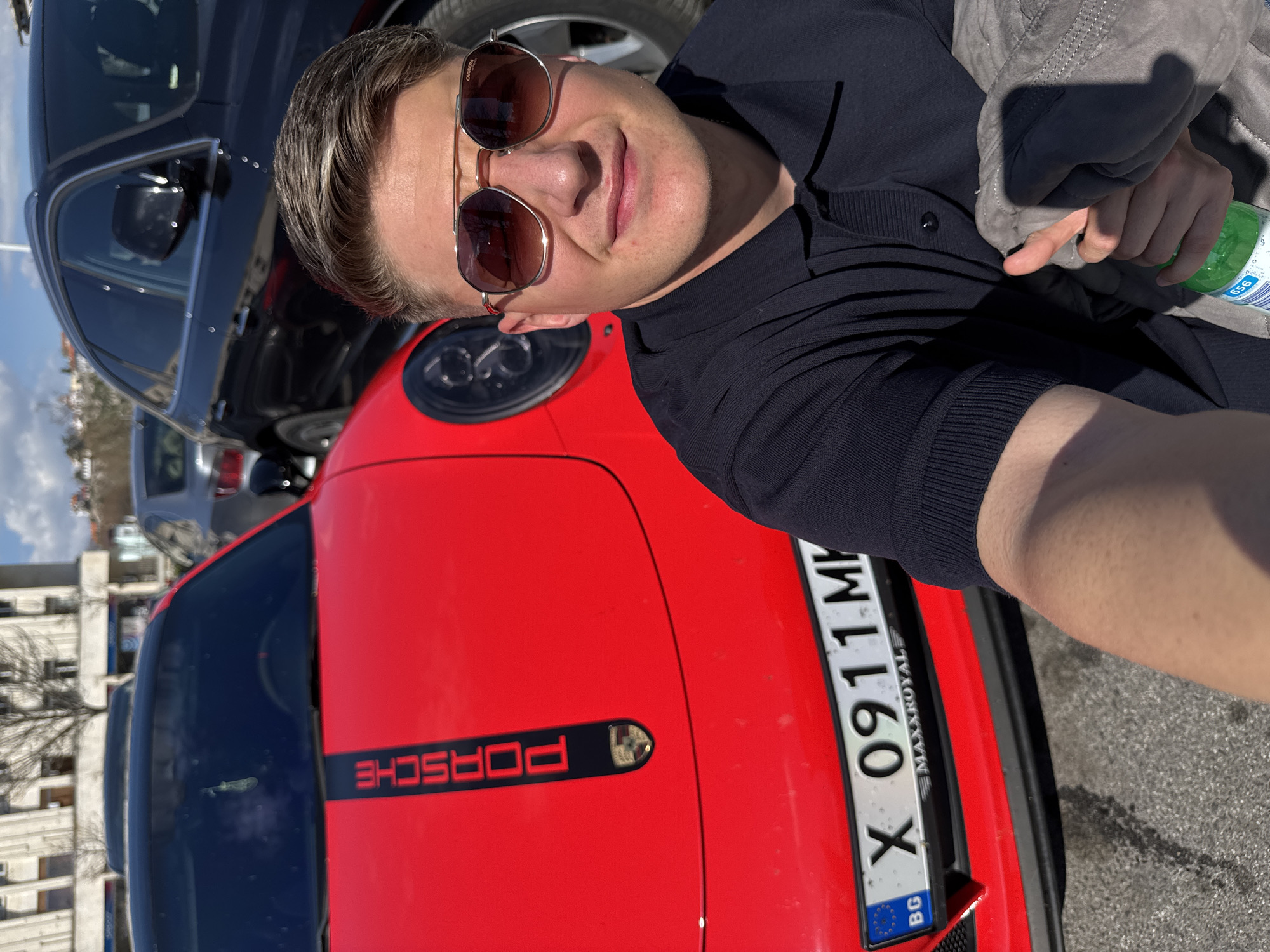 A man is taking a selfie outdoors next to a red Porsche. The Porsche has a black stripe on the hood with the word "PORSCHE" on it. The license plate on the Porsche reads "X 0911 MH BG." The man is wearing sunglasses and a dark-colored shirt, and he is holding a jacket and a bottle of water. Behind the Porsche, there are other cars and buildings visible. The image was taken in Plovdiv, Bulgaria.