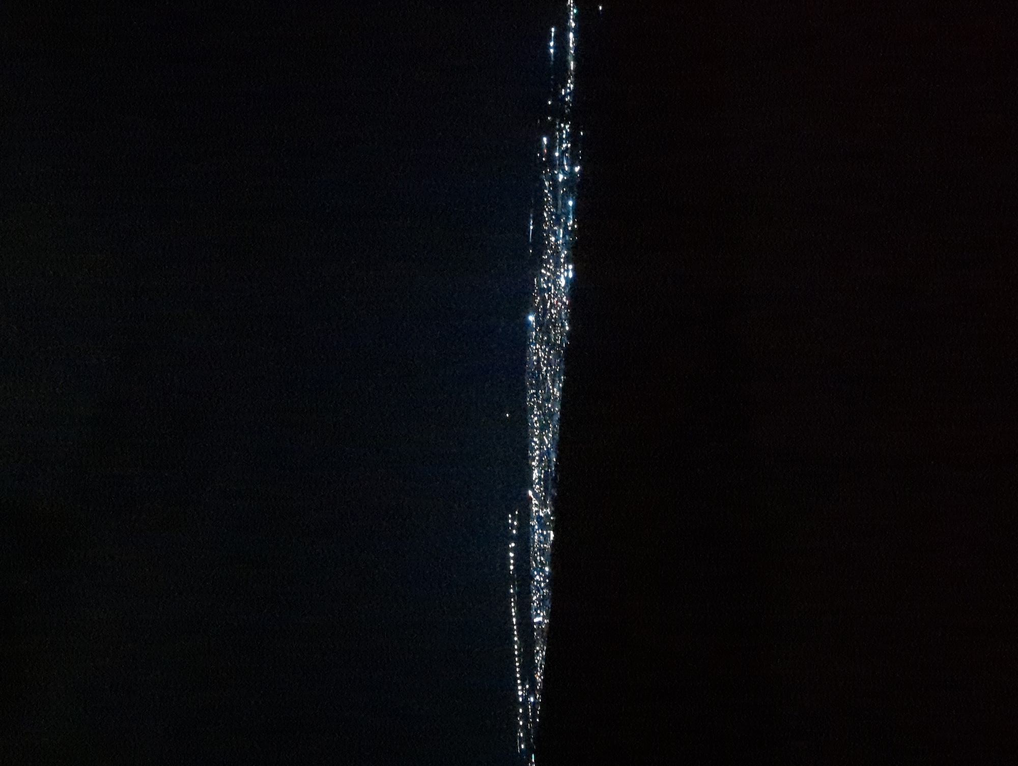 A stream of liquid, possibly water, is visible against a dark background. The liquid appears to be fragmented into droplets, creating a sparkling, reflective effect along its central path. The background is predominantly dark, with minimal texture. There is no indication of a specific location or city, let alone Razgrad, Bulgaria. The image features no people, specific objects, or actions beyond the falling liquid.