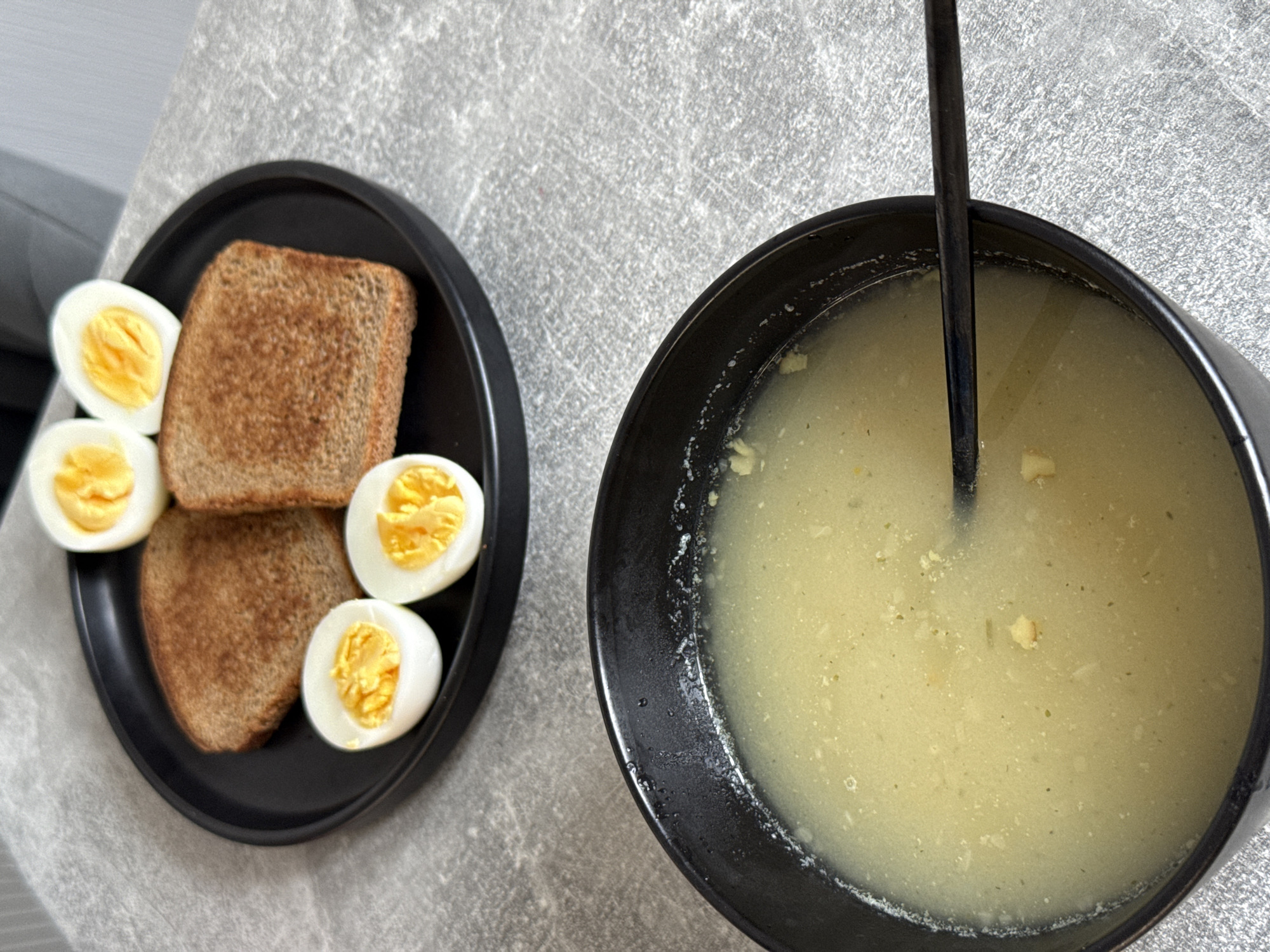 The image shows a breakfast meal laid out on a gray, textured surface. In the foreground, a black bowl contains a light-yellow soup with small solid particles, and a dark utensil rests diagonally within it. In the background, a black plate holds two slices of toasted brown bread and four halves of hard-boiled eggs, arranged around the toast. The eggs have bright yellow yolks. The scene appears to be indoors, likely a kitchen or dining area. There is no indication of the city of Kraskovo, Russia, or any specific location.