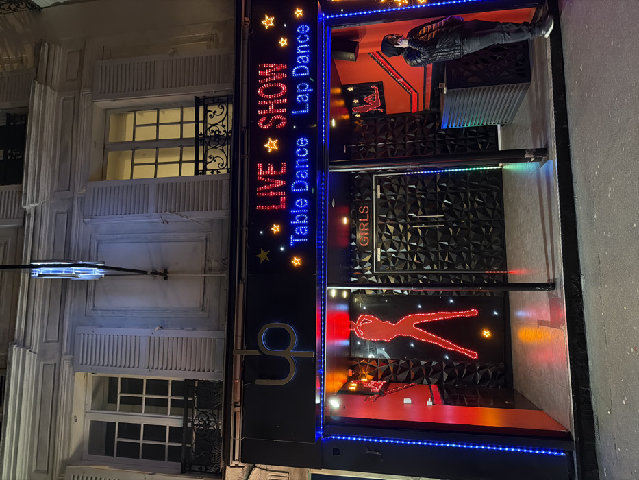 A nighttime street view reveals a business establishment with the signage "LIVE SHOW, Table Dance, Lap Dance," punctuated by star icons. The entrance features a door marked "GIRLS," a red silhouette resembling a dancer, and a black textured wall. A person in dark attire is visible within. The building's façade incorporates traditional architectural elements like shuttered windows and stone detailing.