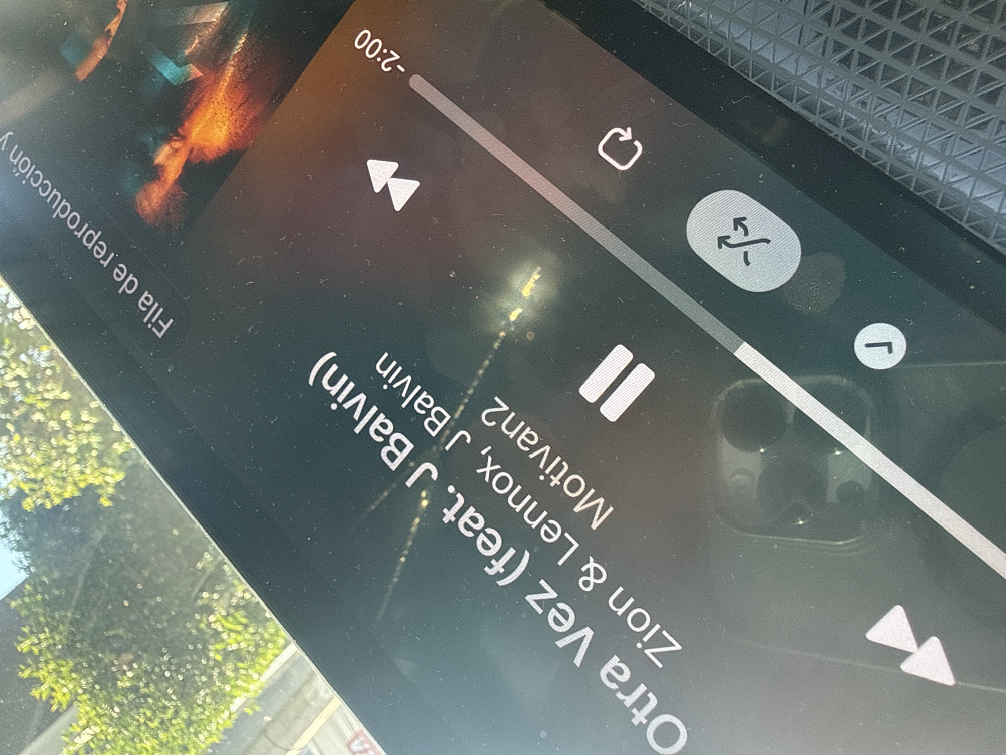 A tilted close-up shot captures a smartphone screen displaying a music player interface. Bright reflections of sunlight and green foliage are visible on the screen, indicating an outdoor or sunlit setting. The screen shows the song "Otra Vez (feat. J Balvin)" by artists "Zion & Lennox, J Balvin," belonging to an album or playlist labeled "Motivan2." The text "Fila de reproducción" (Spanish for "Playback queue") is also visible along the left edge.
The progress bar indicates that approximately "-2:00" minutes remain in the current track. Several playback controls are present: icons for playing the previous track (double left arrows), repeating the current track or playlist (circular arrow), shuffling tracks (crossed arrows), and pausing the music (two vertical lines). The next track icon (double right arrows) is partially visible. Reflections on the screen reveal what appears to be the camera bump of another phone. The background behind the screen features a dark, textured surface, possibly part of a car's dashboard or interior, consistent with the external context of Puebla, Mexico. The screen shows some dust and minor smudges.