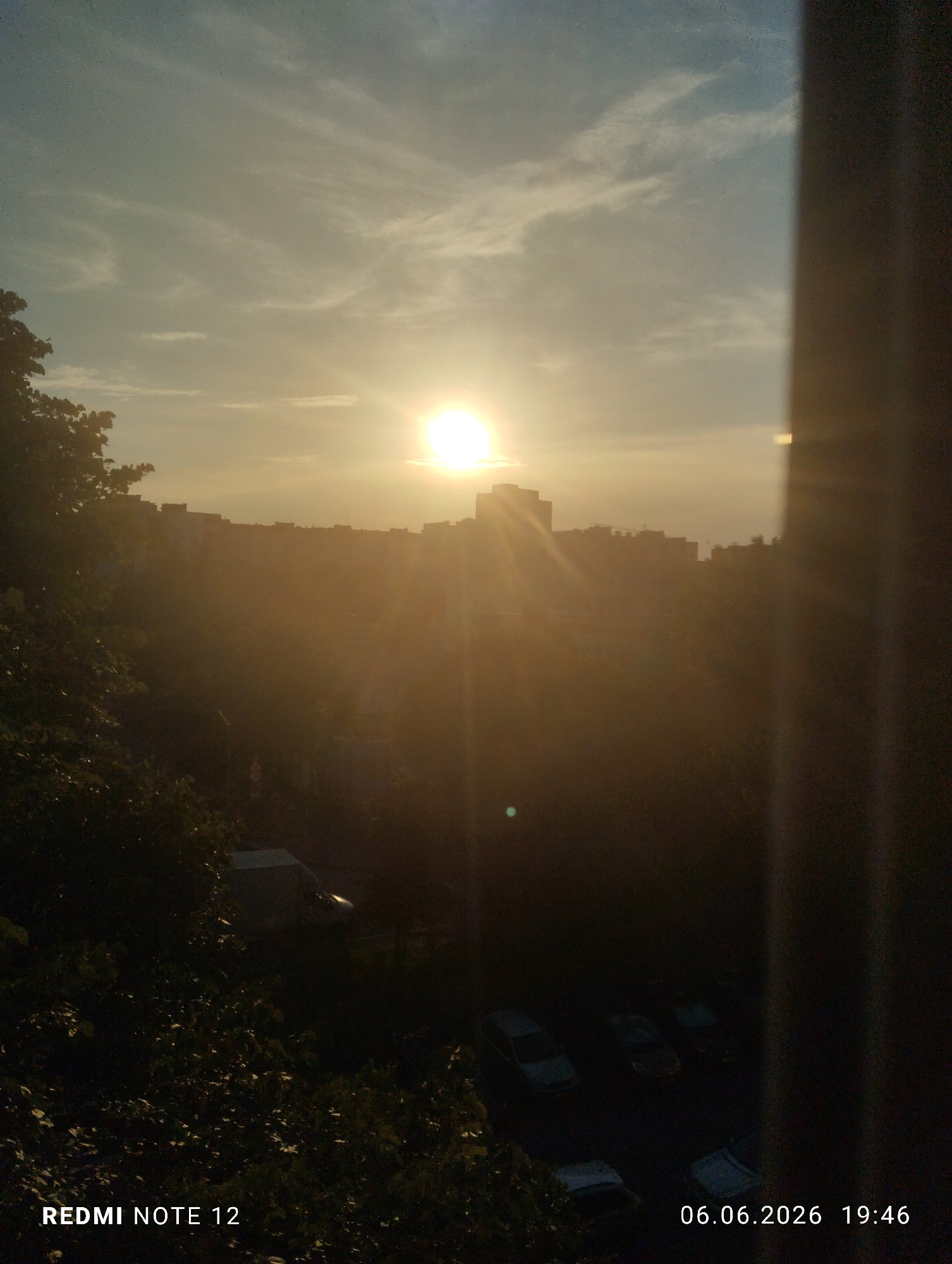 The image depicts a sunset over the city of Burgas, Bulgaria, captured on June 6, 2026, at 19:46, using a Redmi Note 12 device. The sun, a bright yellow-white orb, is positioned in the upper-middle-right of the frame, emitting prominent lens flare rays that extend across the central portion of the scene. The sky transitions from a light blue with sparse white clouds at the top to a bright yellow-orange near the horizon, indicative of late afternoon conditions.
A silhouette of multi-story buildings forms the city skyline, heavily backlit and partially obscured by the intense sunlight and atmospheric haze. A construction crane is visible atop one of these structures in the middle-right. The mid-ground and foreground are significantly underexposed due to the strong backlight. Dense, dark green foliage occupies the entire left side of the frame, extending upwards and obscuring elements below.
A dark, vertical, rectangular element, consistent with a window frame or similar obstruction, runs along the far right edge of the image, partially blocking the view. In the heavily shadowed bottom section of the image, several indistinct light-colored vehicles are faintly discernible in what appears to be a parking area or street. A white van-like vehicle is partially visible further left, obscured by foliage. A small, faint, circular, blue-green light artifact is present in the lower-middle-left quadrant. No individuals are visibly present in the frame.