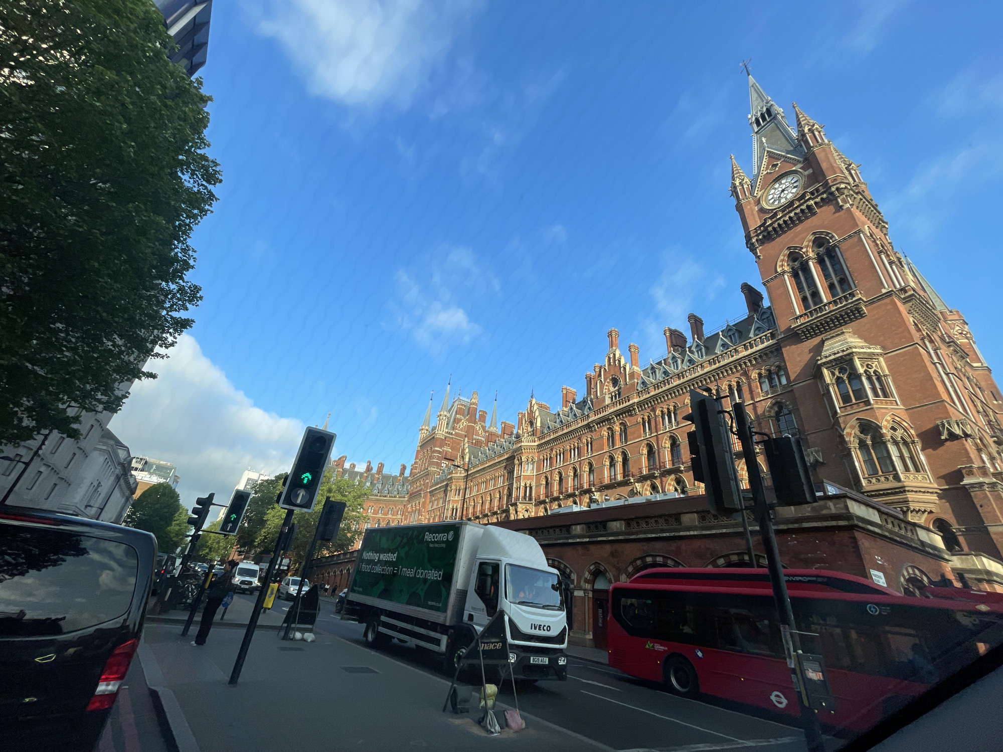 The image captures a street scene in London, United Kingdom, featuring the iconic St. Pancras Renaissance Hotel, distinguishable by its Victorian Gothic architecture and prominent clock tower. A white Iveco truck with "Recorra" and the slogan "Nothing wasted, 1 food collection = 1 meal donated" is in the street. Beside the truck is a street sweeping station. A red double-decker bus is partially visible to the right of the truck. Several traffic light poles are visible, with one showing a green light indicating "go." A black van is on the left side of the frame, with pedestrians on the sidewalk. The sky is blue with scattered clouds. The overall environment appears urban and bustling.