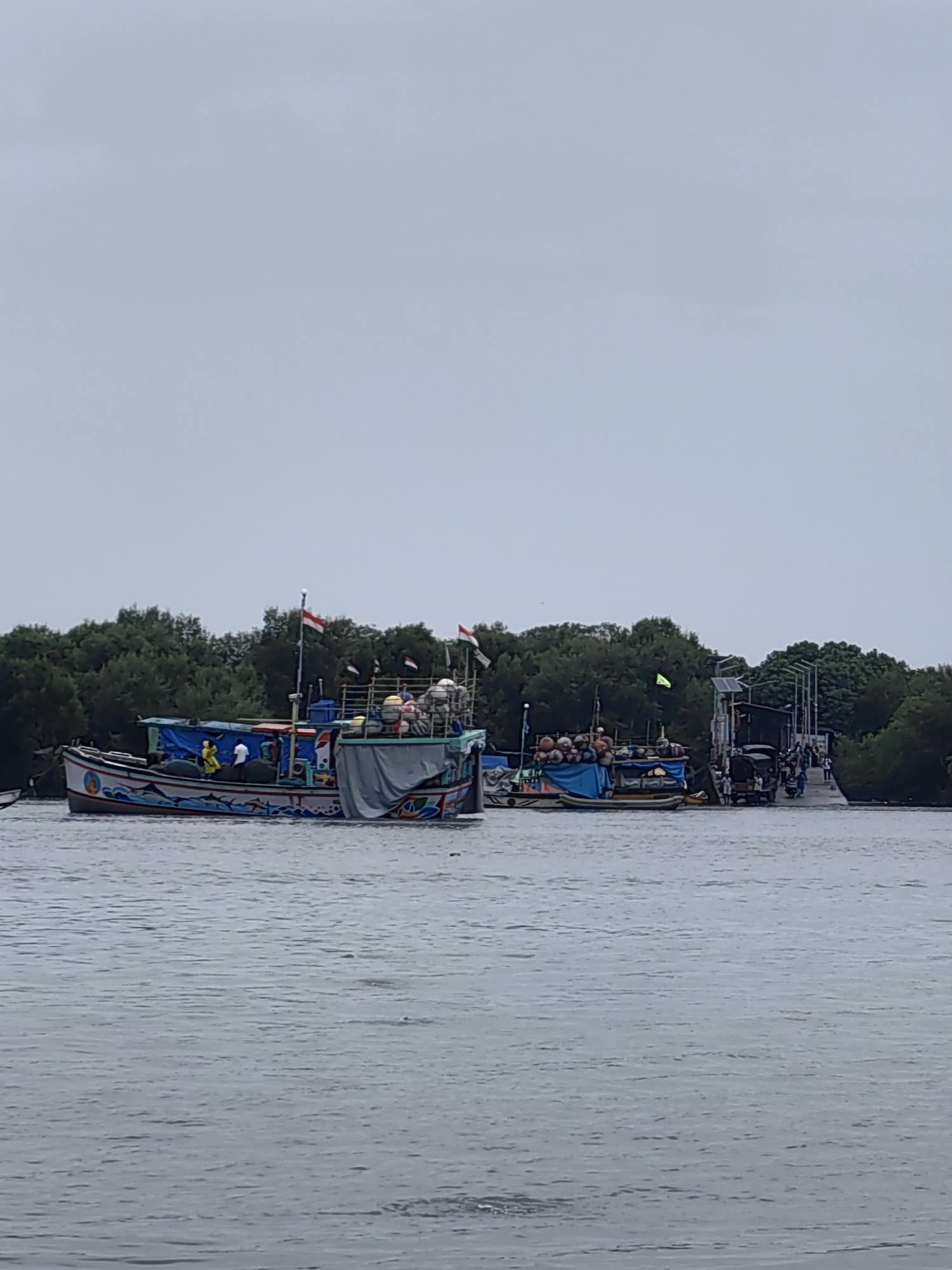 This is a photograph of boats on the water near Gorai, India. The sky is overcast and the water is choppy. In the foreground, there are two fishing boats. The boat on the left is larger and painted with colorful designs. It has several people on board. The boat on the right is smaller and appears to be loaded with supplies. In the background, there is a dense line of trees and a structure that looks like a pier or a bridge. Several people are visible on this structure, and there are also some vehicles, including a tuk-tuk and motorcycles. The overall scene suggests a busy harbor or fishing village.